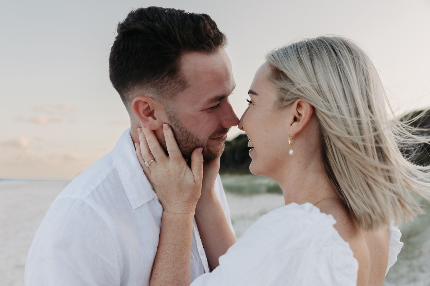 A romantic couple in white clothing sharing an intimate moment at sunset by the water.