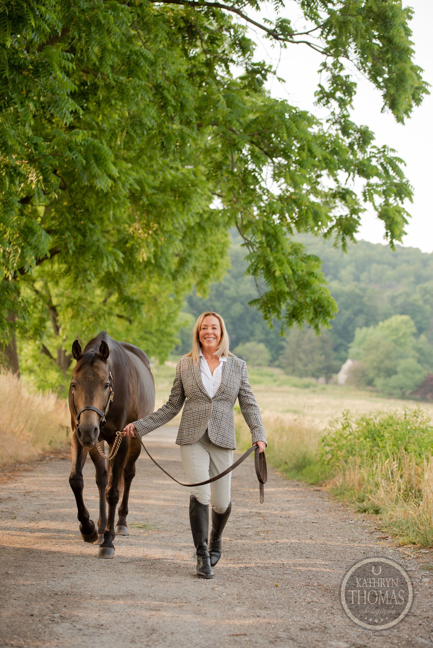 Handler in riding attire leads a bay horse down a scenic wooded trail.