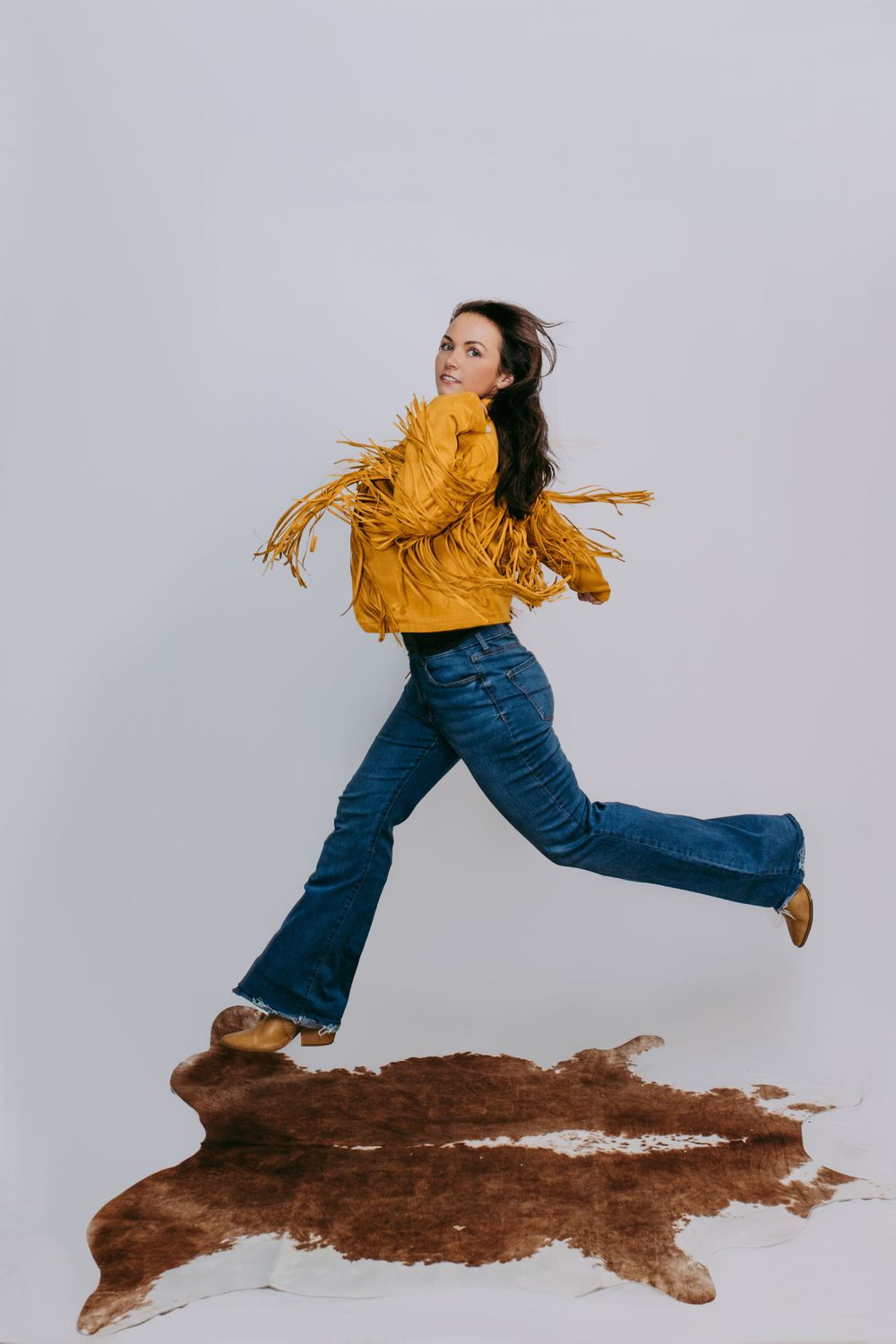 A dynamic figure in a yellow jacket and flared jeans appears to be floating over a brown cowhide rug against a white background.
