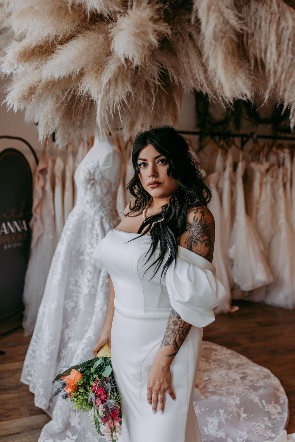 Modern bride in off-shoulder white dress with tattoos poses against backdrop of pampas grass and wedding gowns.