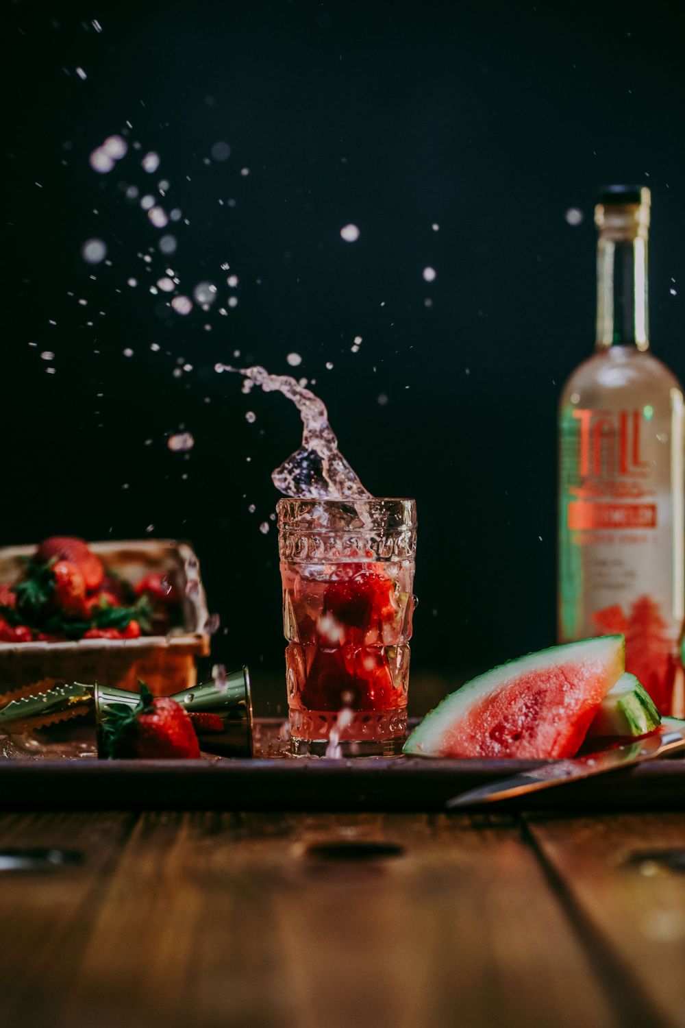 Red cocktail splash with watermelon and Tall Pines bottle on rustic wooden table.