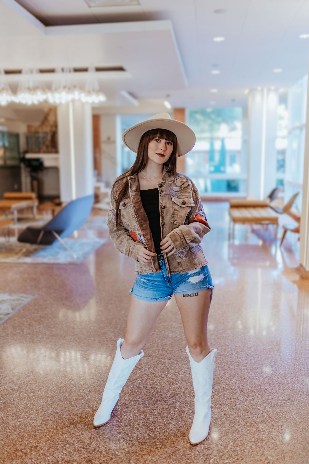 A person poses in a cafe wearing a brown jacket, denim shorts, white cowboy boots and wide-brimmed hat.