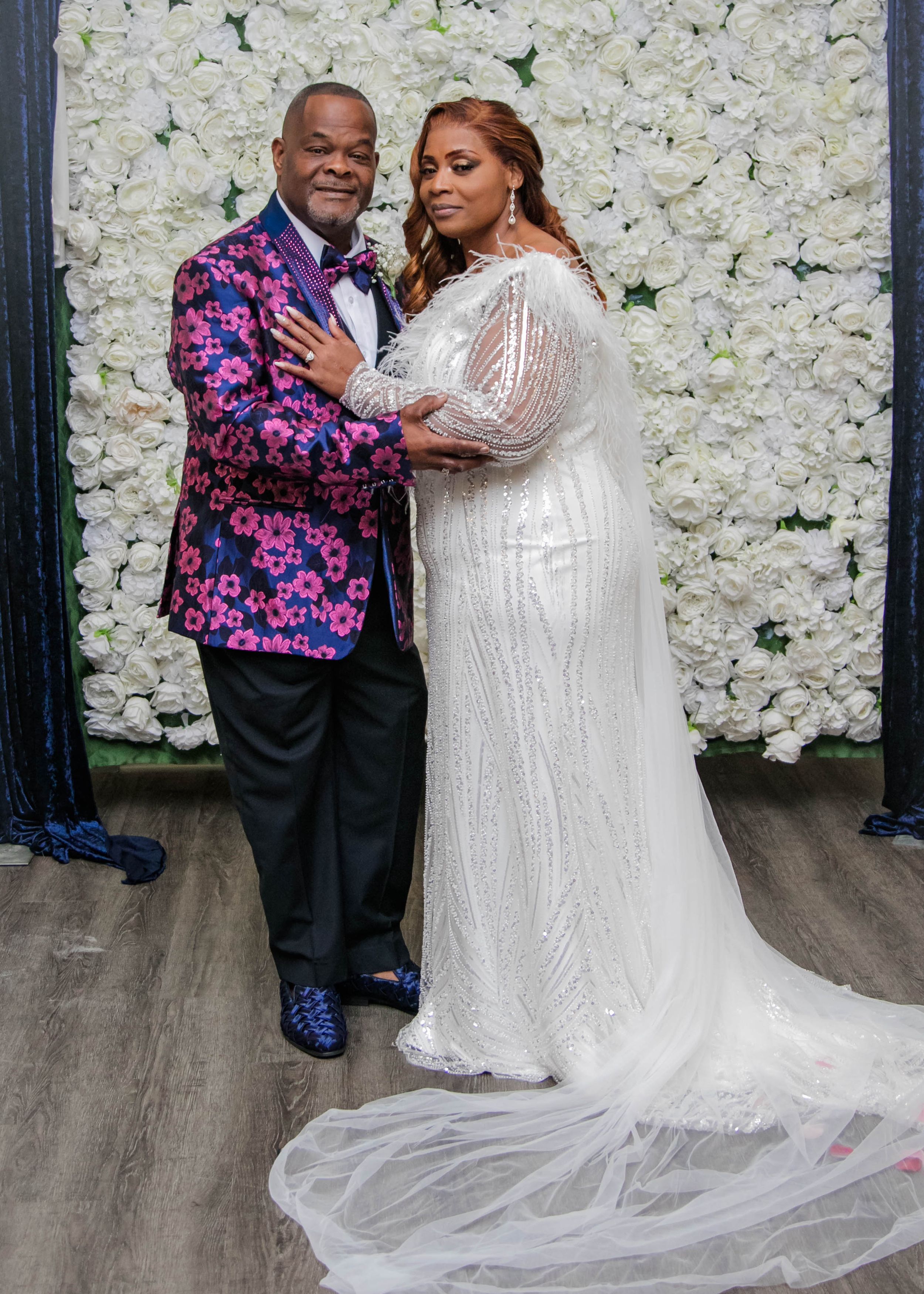 Newlyweds pose against floral wall, with groom in floral jacket and bride in sparkly white gown.