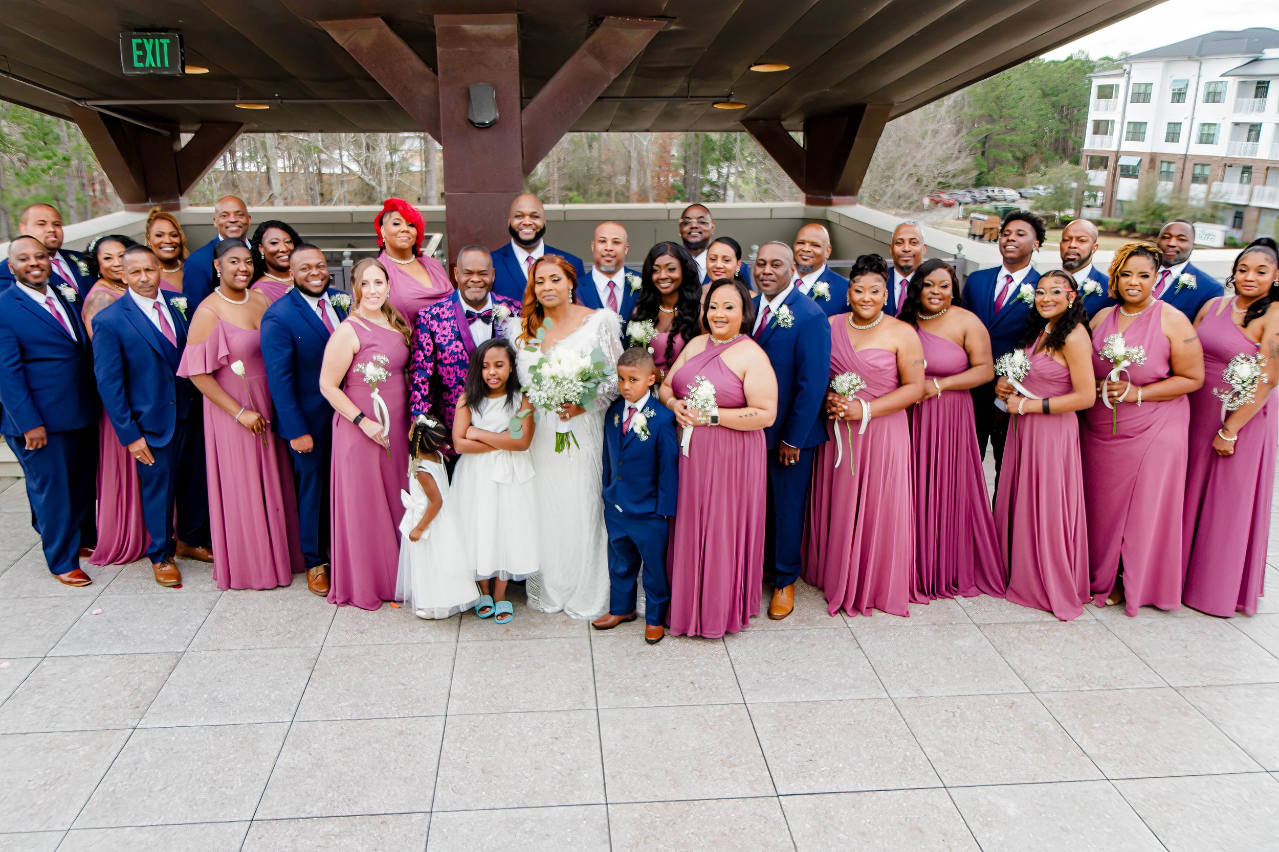 Large wedding party in blue and pink attire poses together under outdoor pavilion.