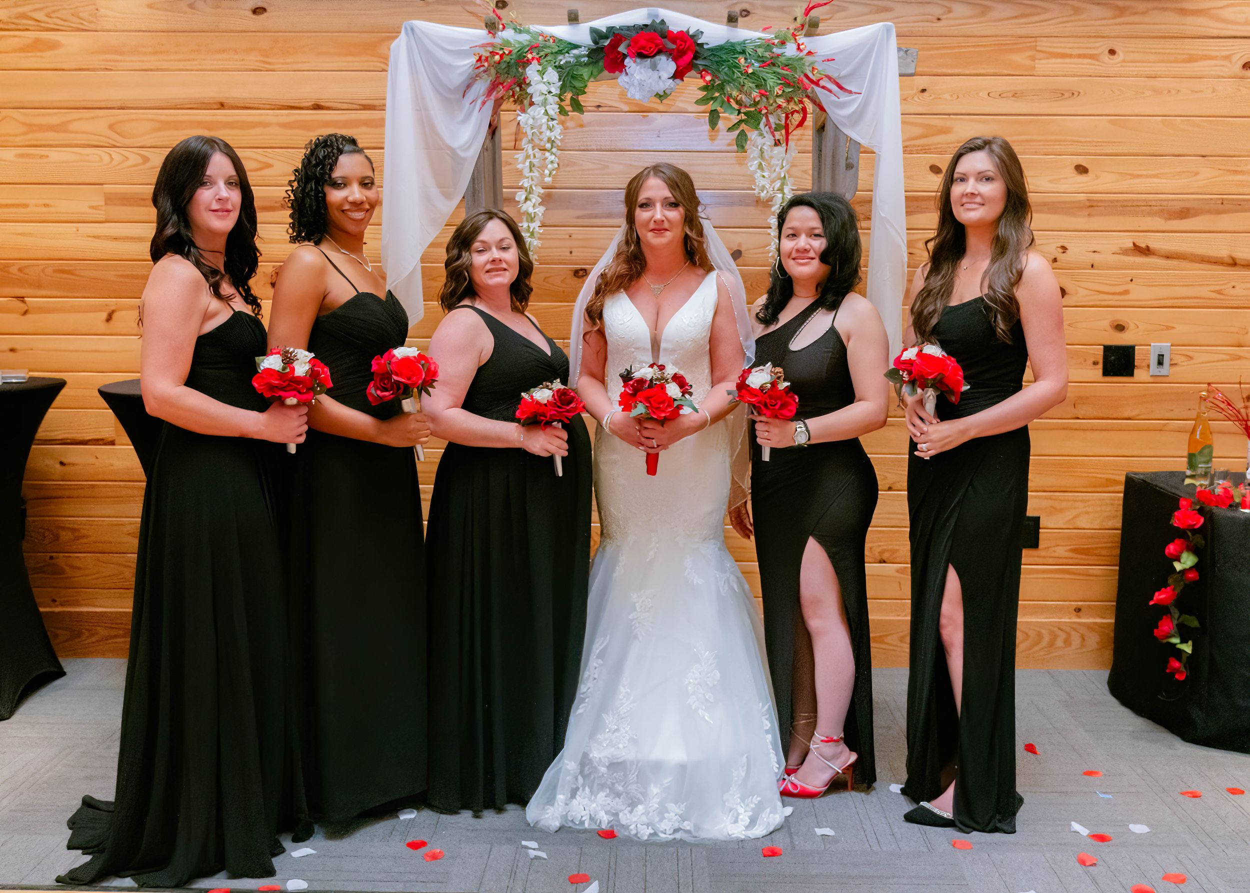 A bridal party in black dresses poses with red bouquets against a wooden wall.
