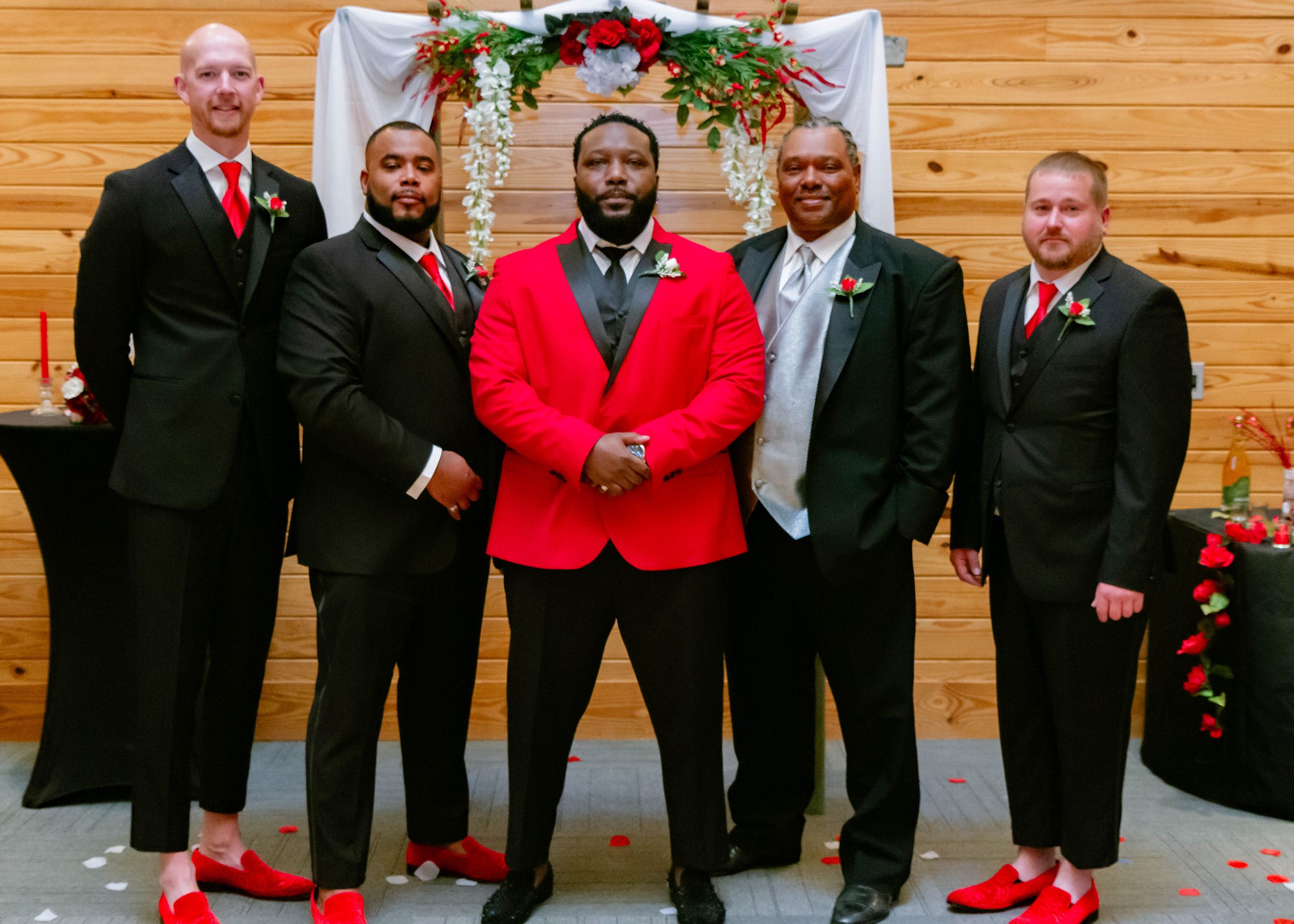 Groomsmen in black suits and red accents pose under a floral arch at a wedding ceremony.