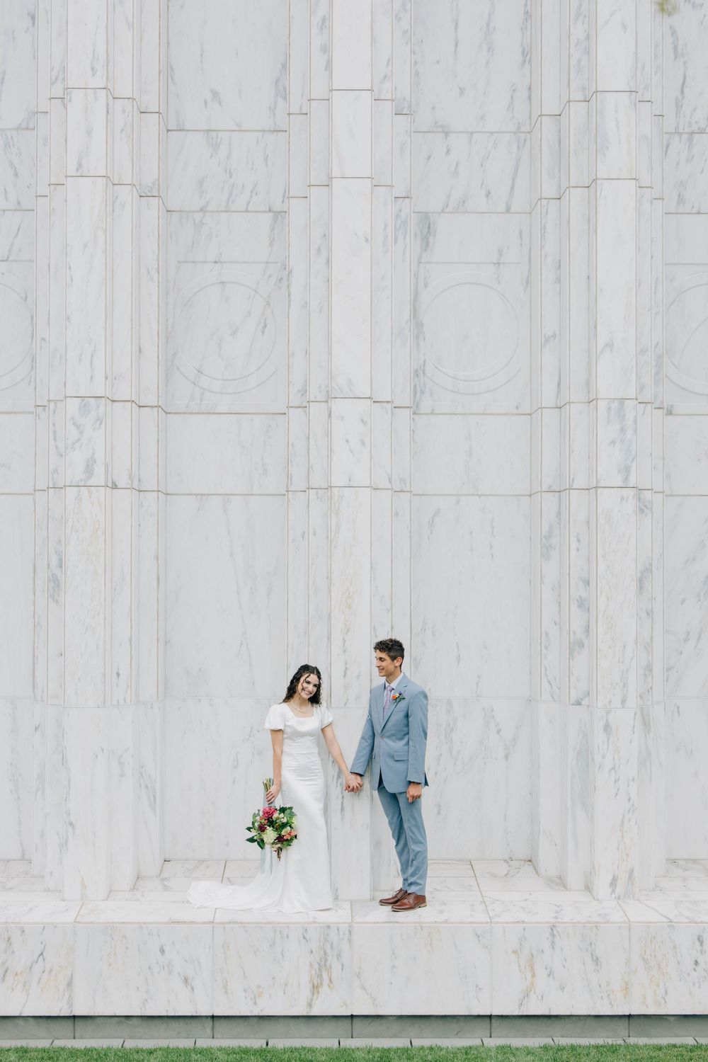 Bride and groom standing apart on marble steps holding colorful bouquet against stone wall.