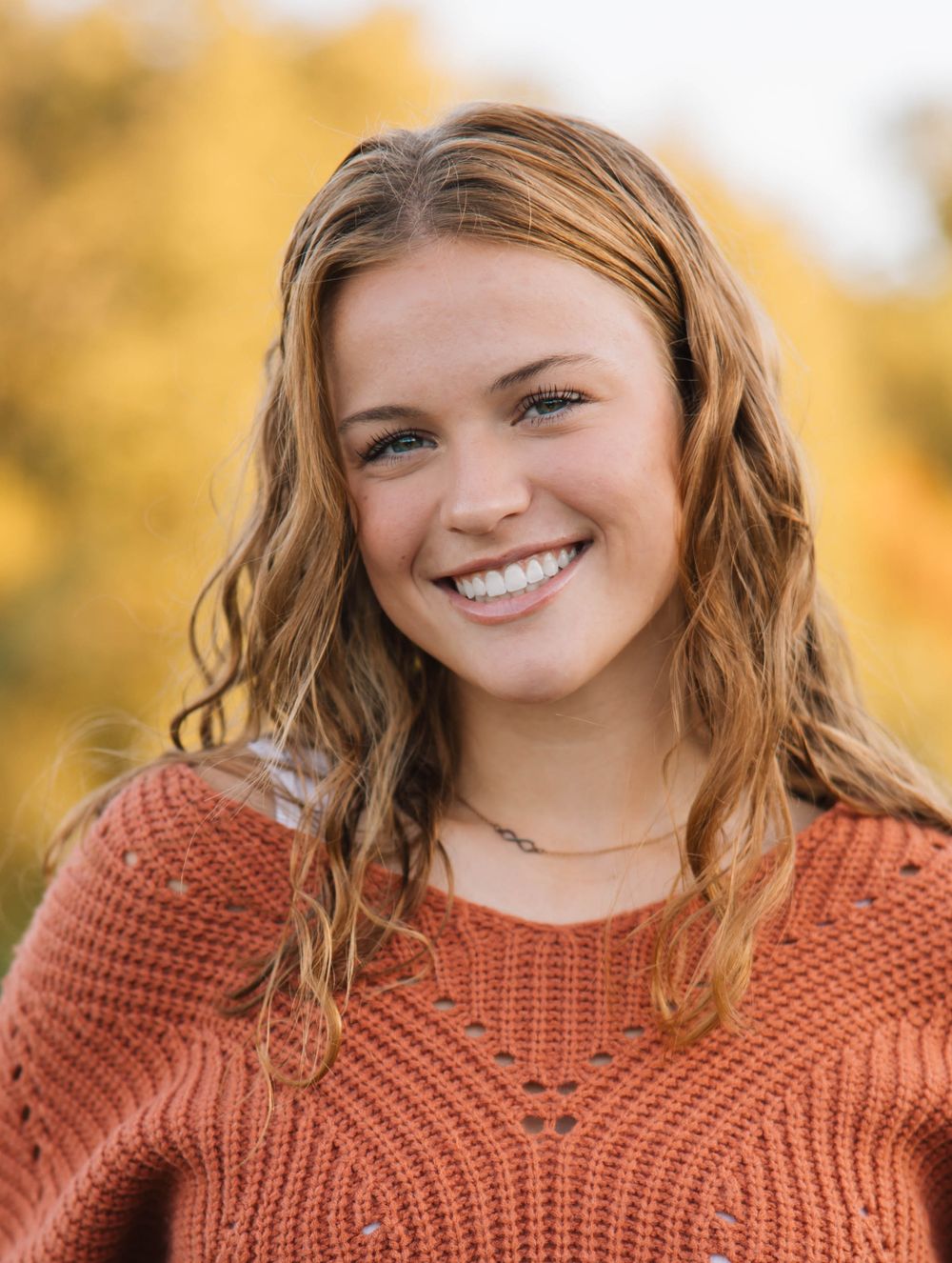 Young woman with blonde hair smiling outdoors in an orange knit sweater with autumn foliage background.