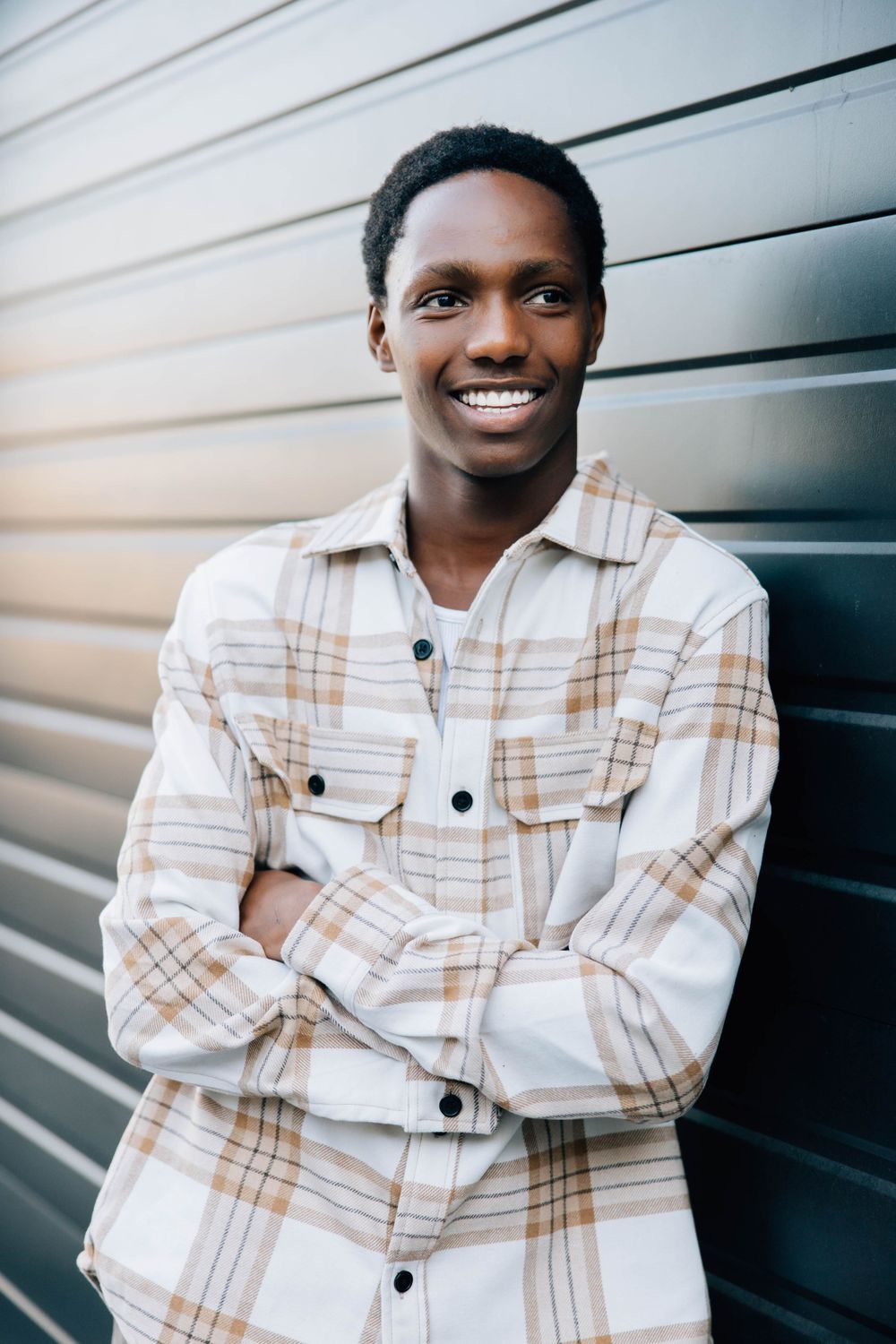 Smiling young Black man with arms crossed wearing a plaid shirt leaning against a metal wall.