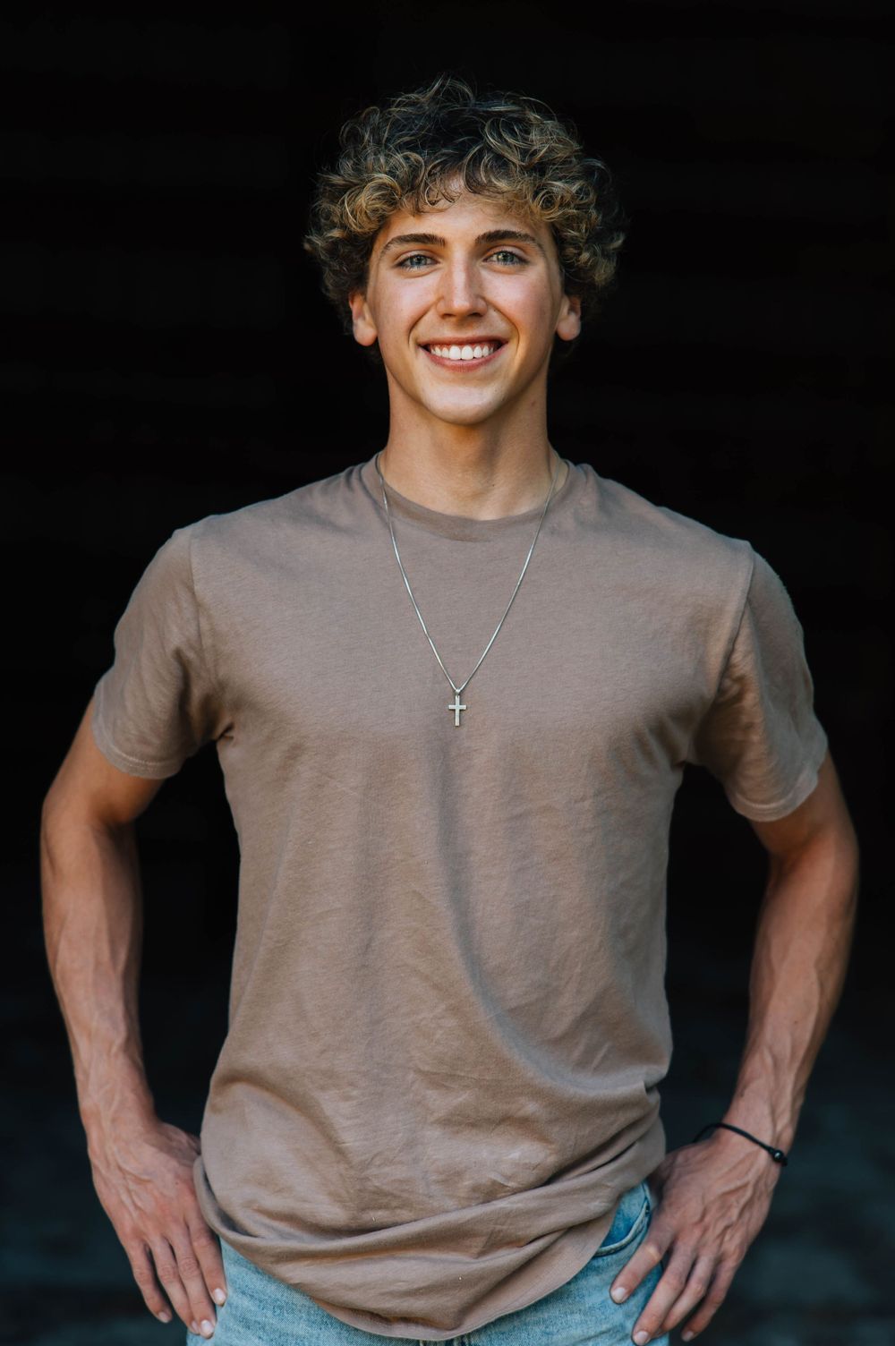 Smiling young man with curly hair wearing a tan t-shirt and cross necklace against a dark background.
