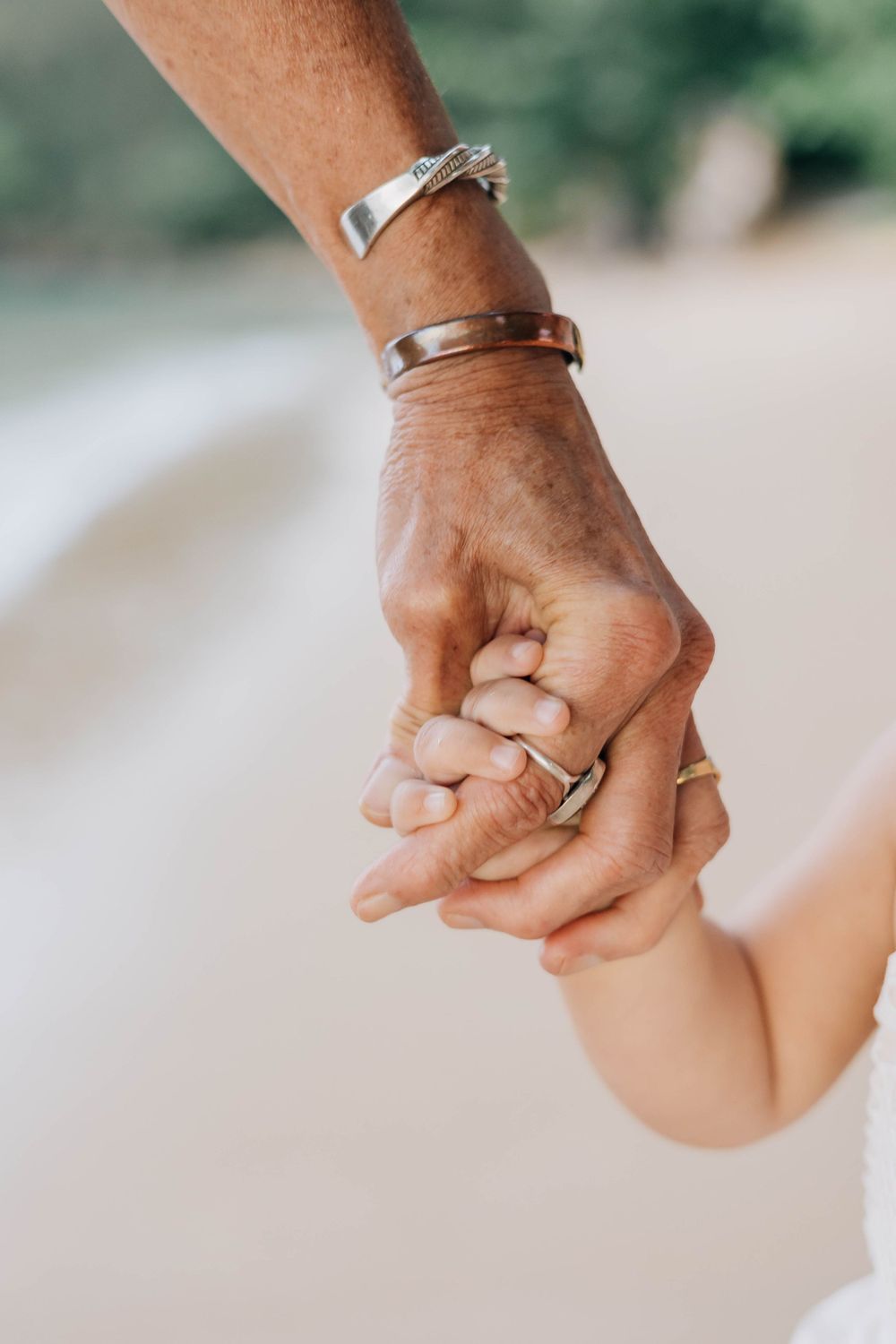 Close-up of adult and baby hands holding together on beach, wearing silver bracelets and rings.