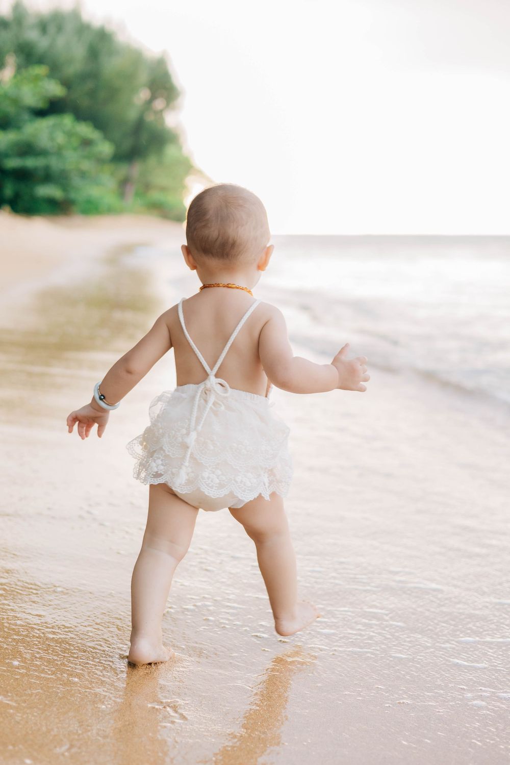 Baby girl in white lace romper walking barefoot on sandy beach shore with ocean waves.