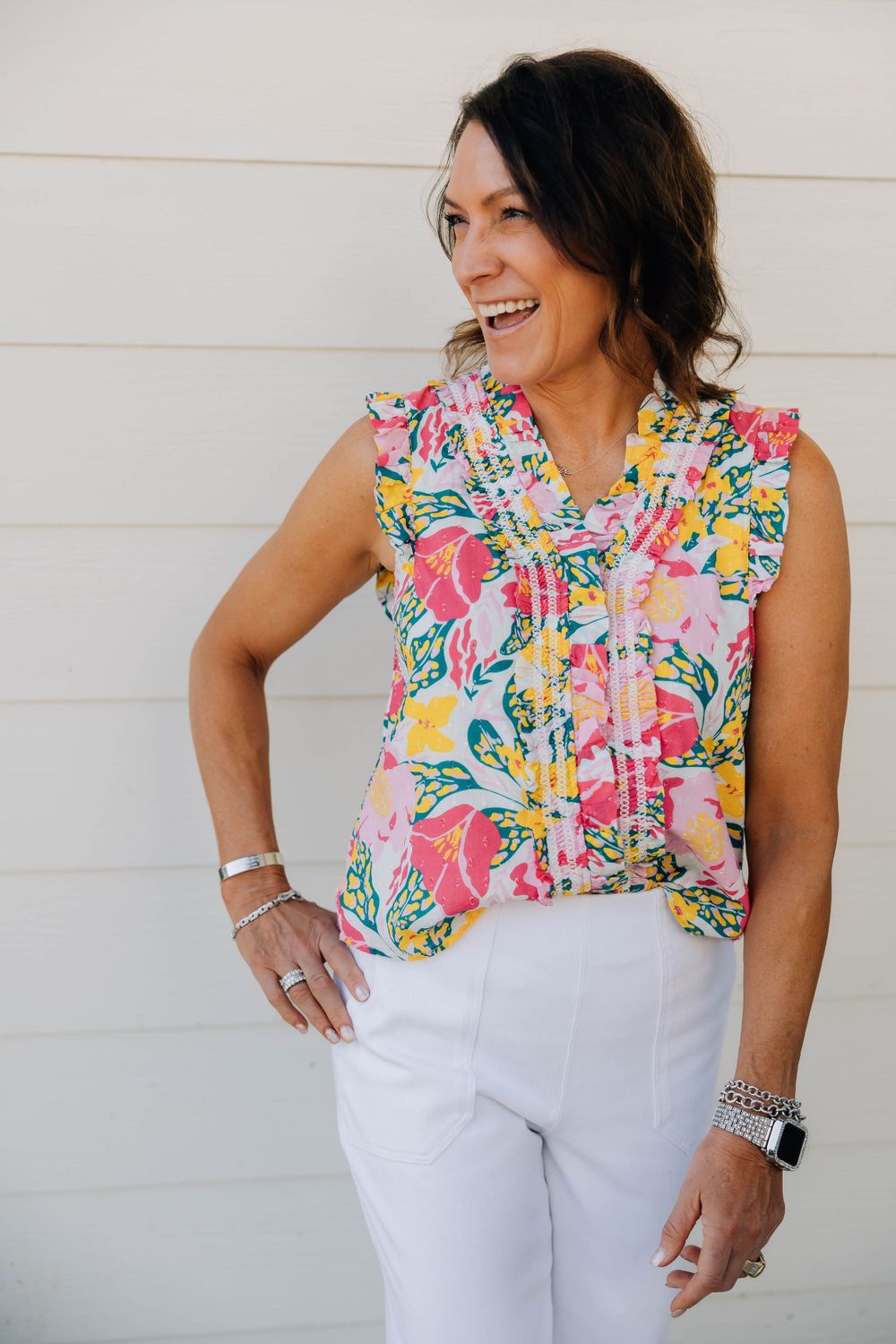 Woman laughing in a colorful floral sleeveless top and white pants standing against a white wall.