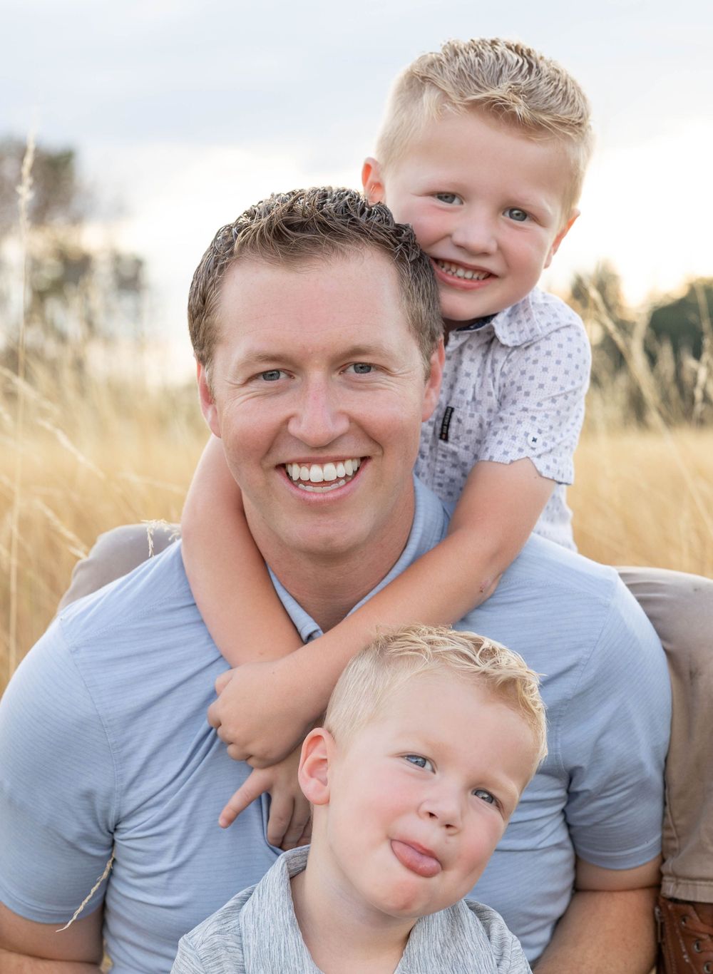 Father smiling with two young blonde boys in golden outdoor grass field.