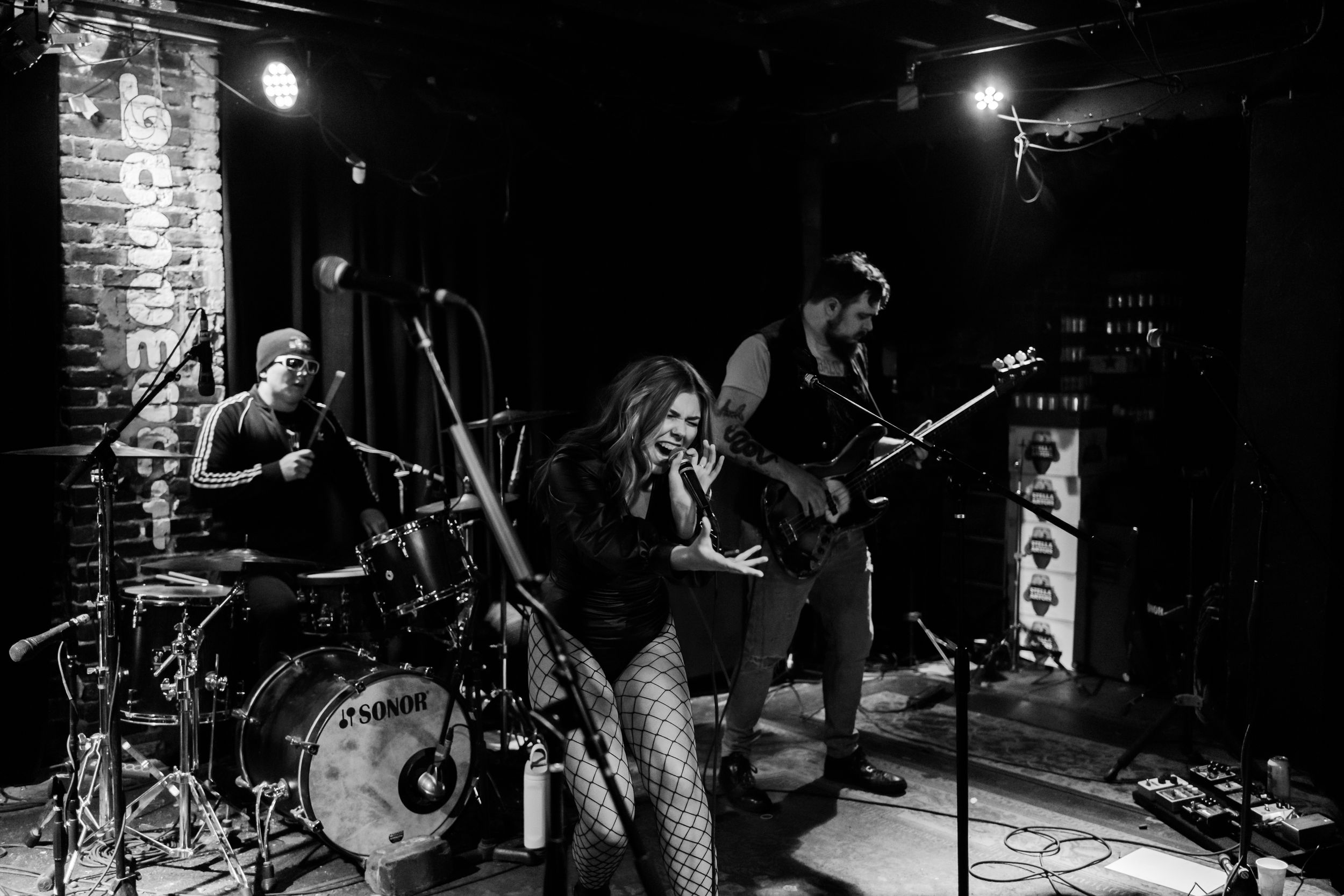 A rock band performs on a small club stage with brick walls and dark lighting.