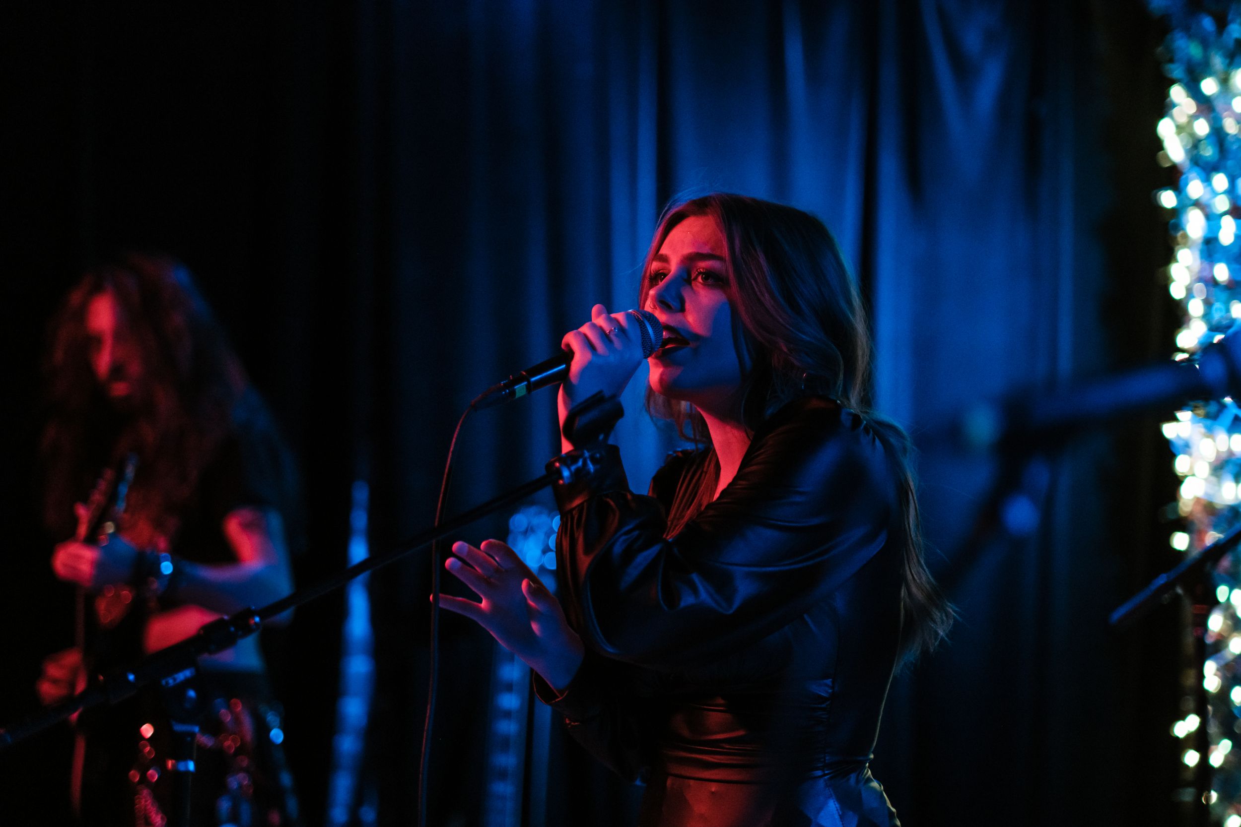 A band performs on stage with blue curtains and string lights creating moody atmospheric lighting.