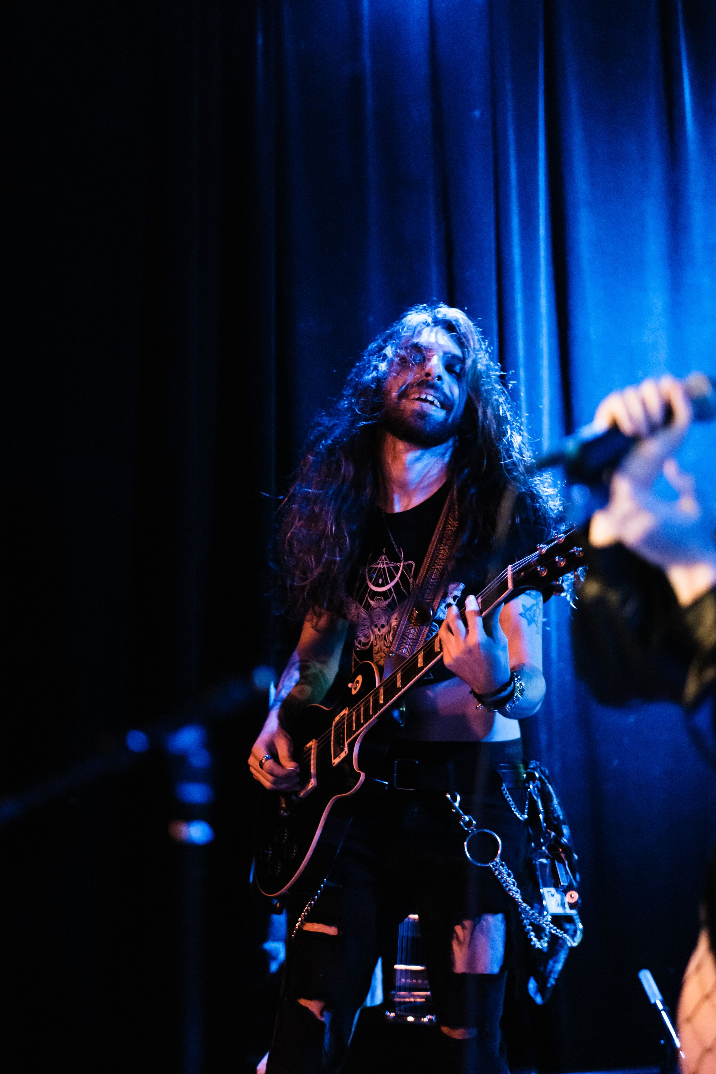 Rock musician with long hair plays guitar under blue stage lighting during performance.
