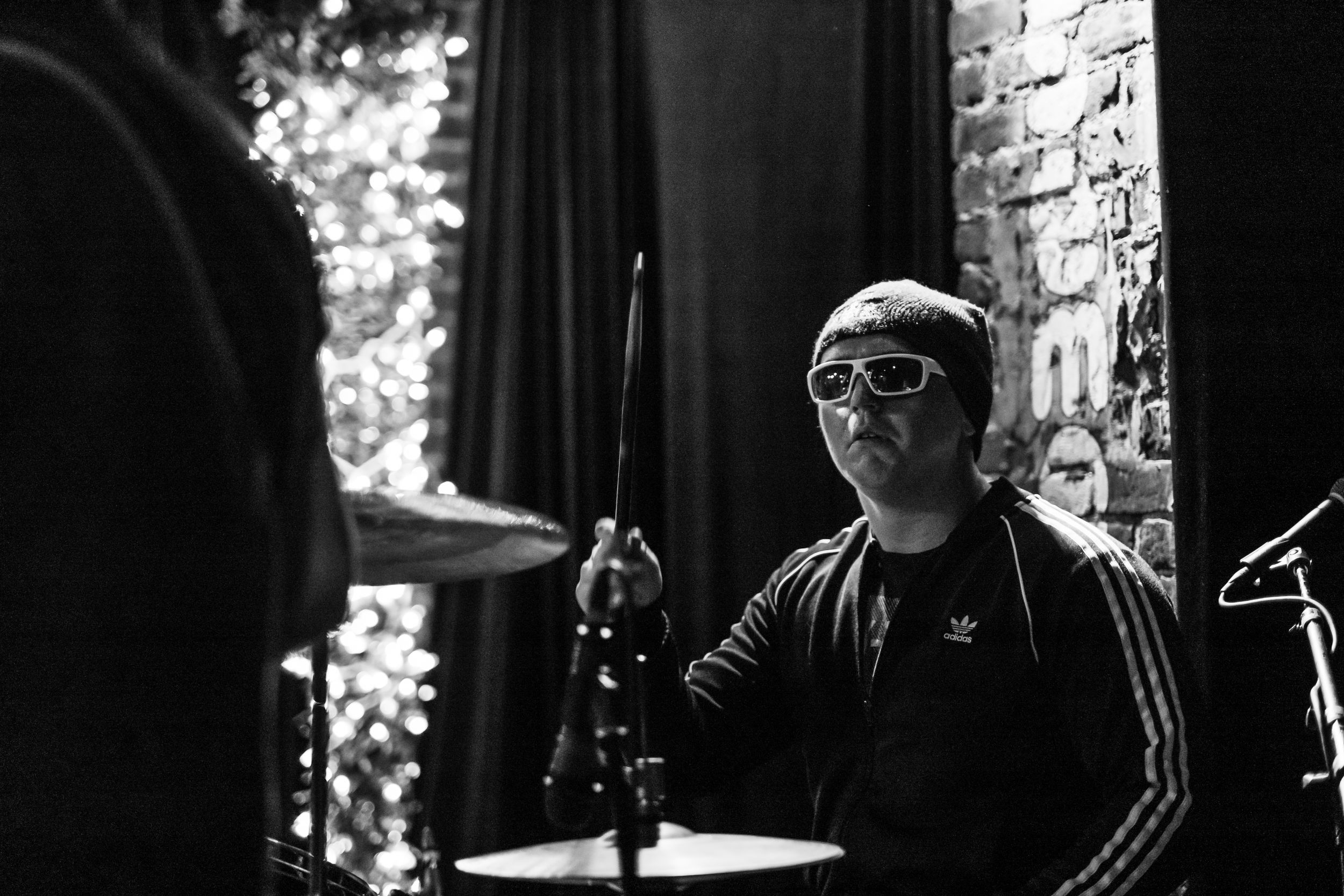 Drummer in black shirt and sunglasses performs during dark concert setting.