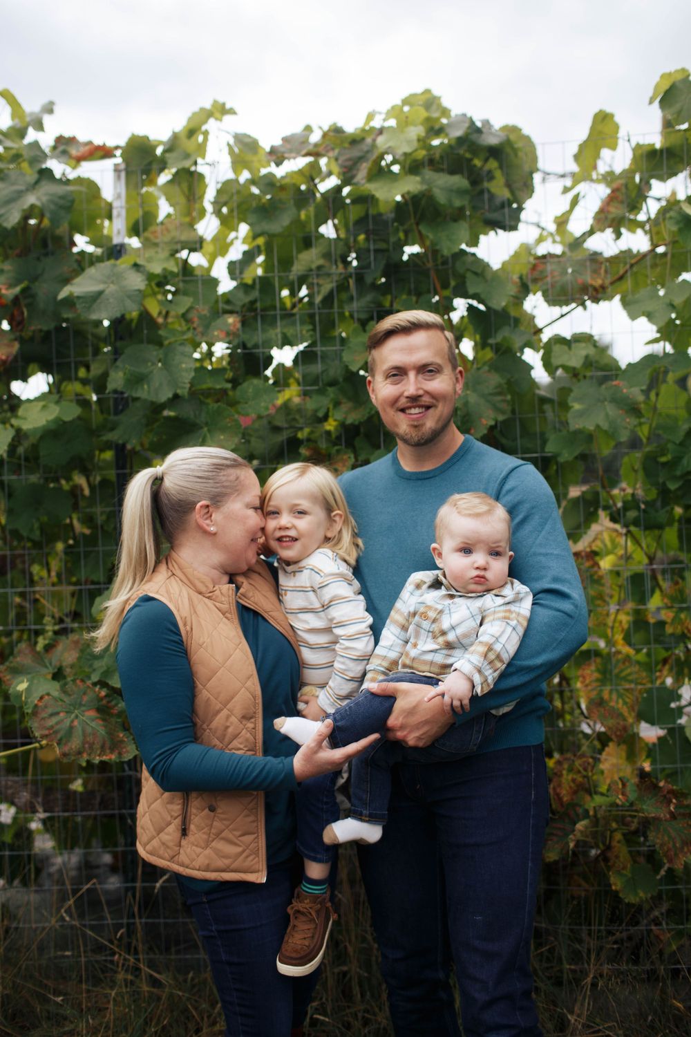 A mother and father carry their newborn and 3-year-old in front of a grapevine at a family photo session in Snohomish