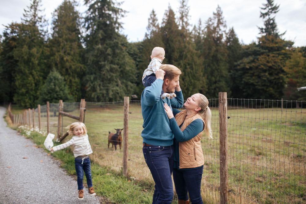A mother and father stand facing each other with a baby on his shoulders. Their young son is skipping in the background