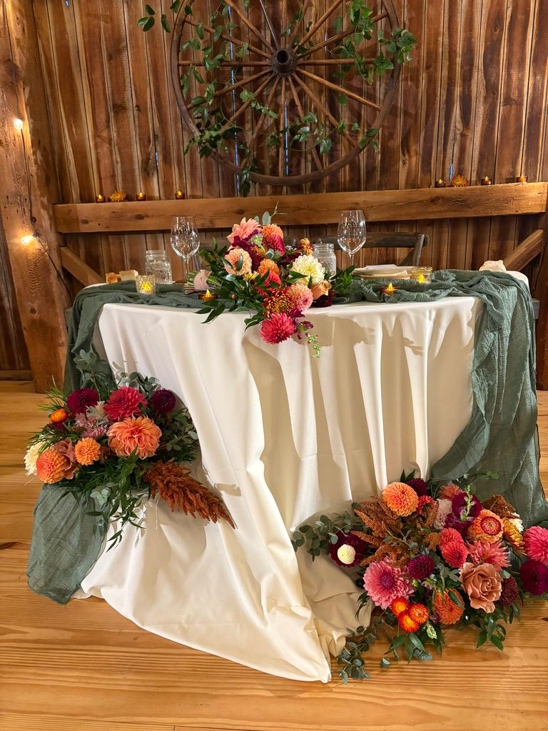 Sweetheart table with white linen and vibrant dahlia floral arrangements in rustic barn reception.