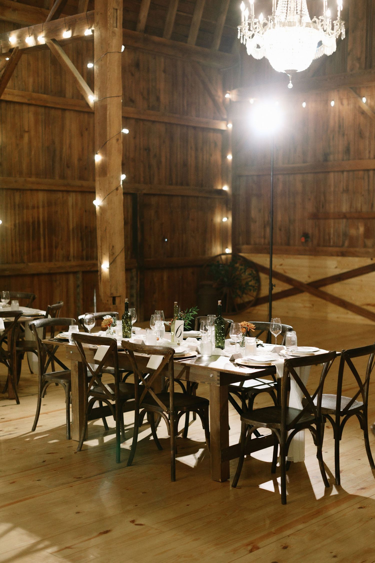 Rustic barn wedding reception with dark wood cross-back chairs, farm tables, string lights, and a crystal chandelier.