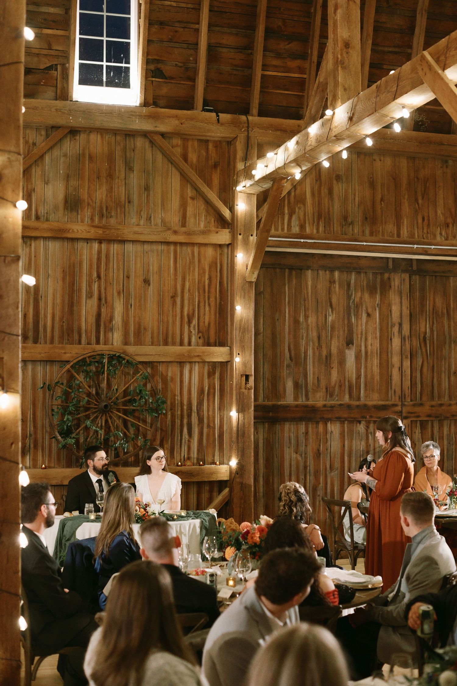 Rustic barn wedding reception with string lights, wooden walls, and guests seated at decorated tables during a toast.
