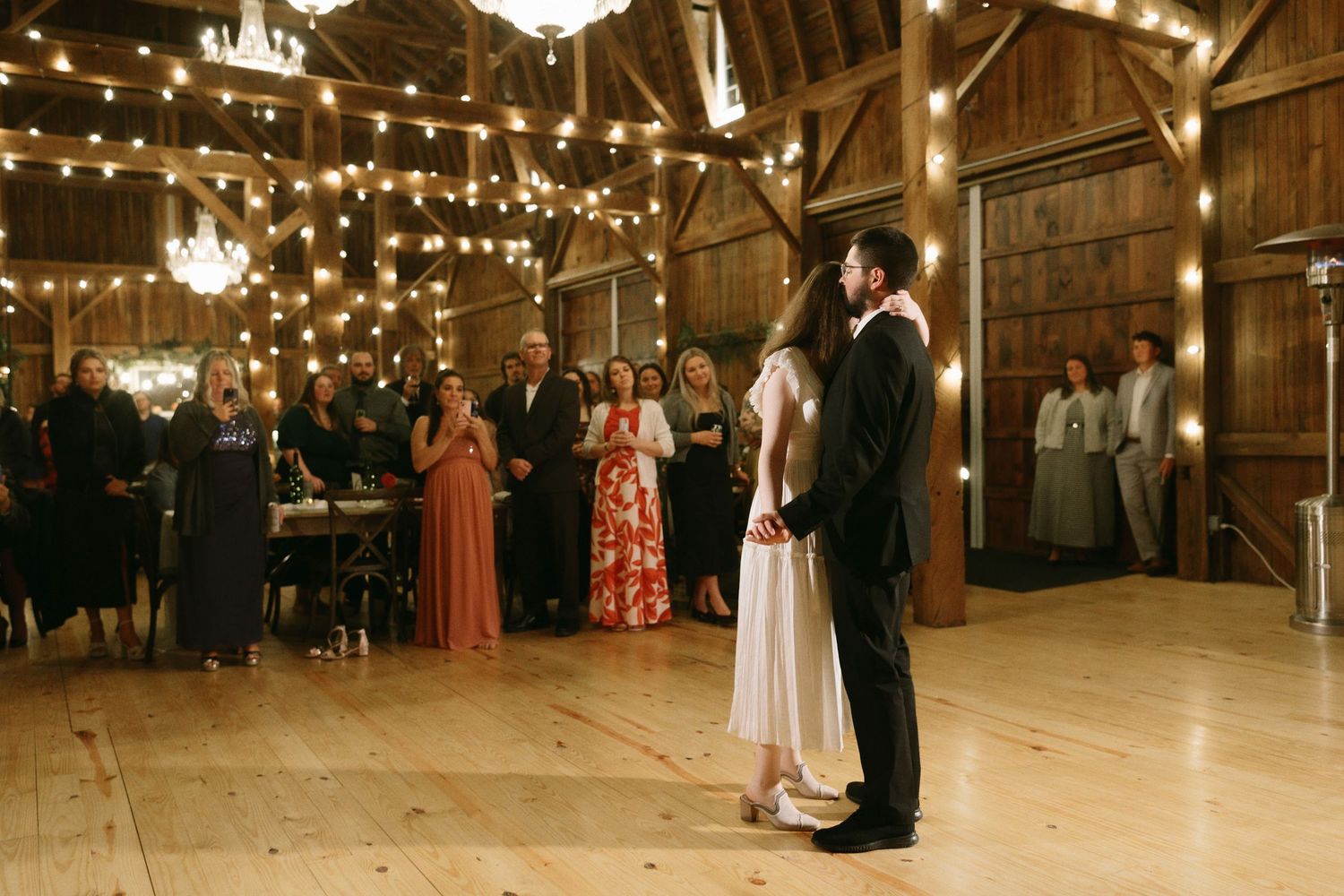 Couple sharing their first dance in a warmly lit rustic barn venue adorned with string lights and wedding guests watching.