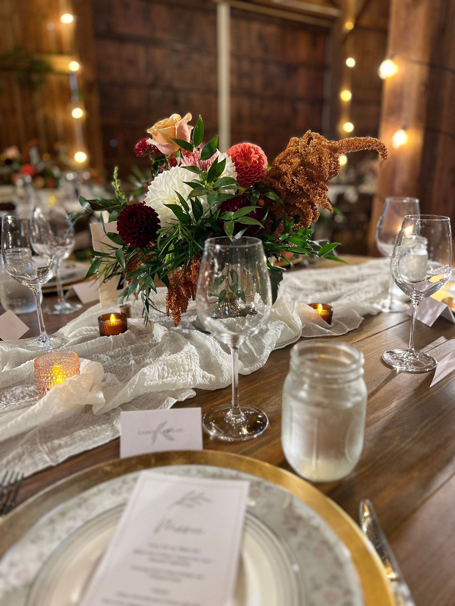 Elegant wedding table centerpiece featuring burgundy dahlias, white flowers, and greenery on a lace runner with gold-rimmed plates.