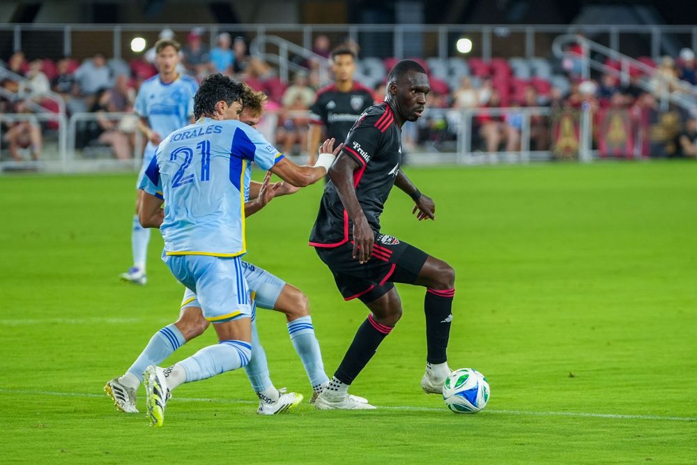 Soccer player in a light blue jersey challenges opponent in a red and black striped uniform during match action.