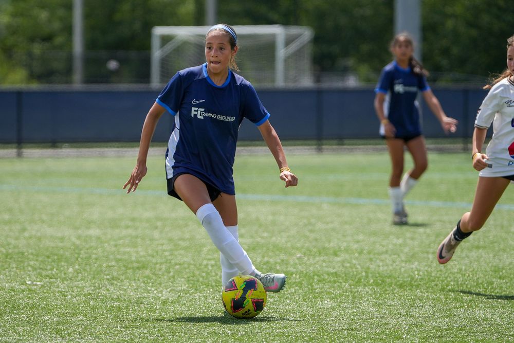 Youth soccer player in blue jersey dribbles soccer ball on turf field during competitive match.