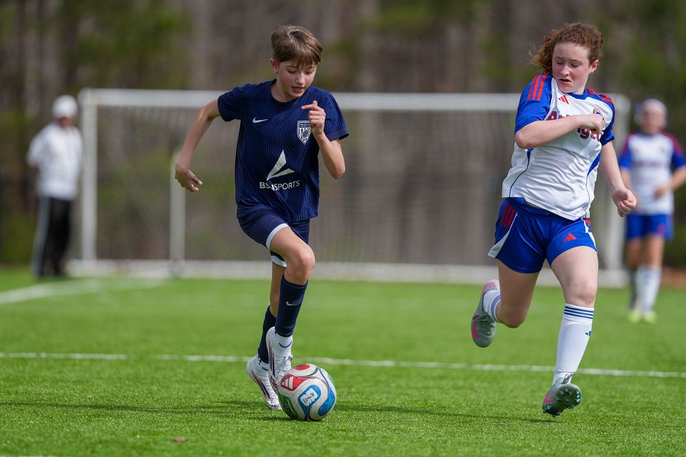 Two youth soccer players compete for the ball during a match on a green turf field.