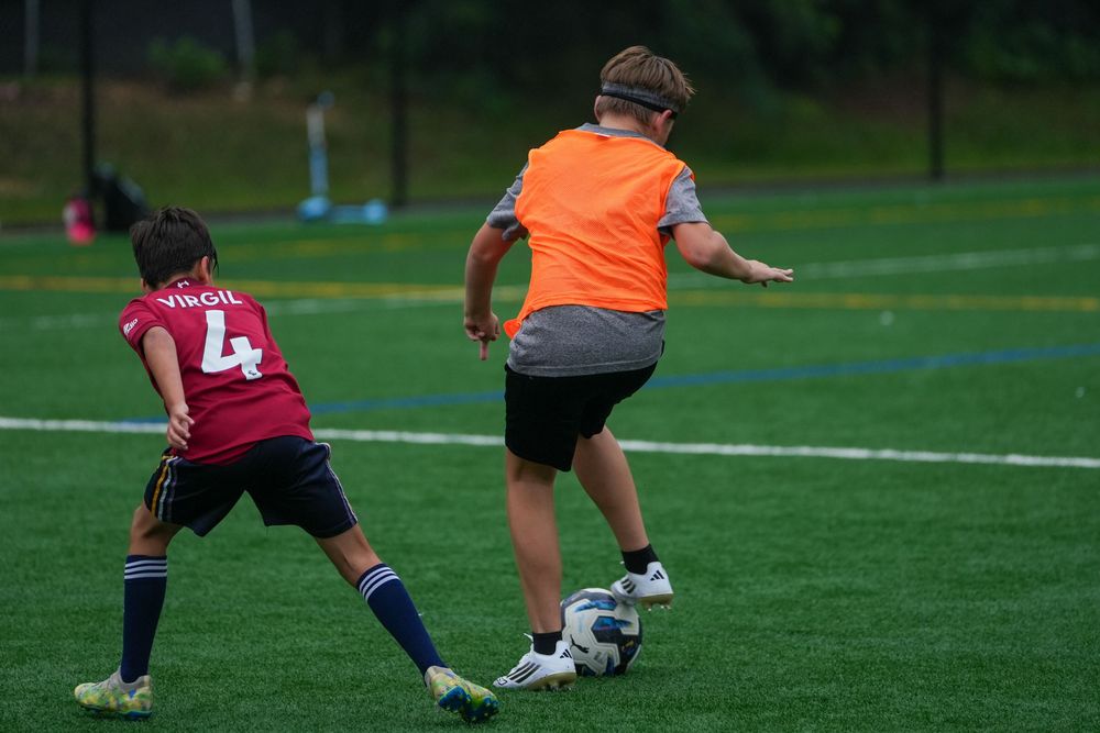 A soccer player in orange jersey shields the ball from a defender in maroon jersey during a match on a grass field.