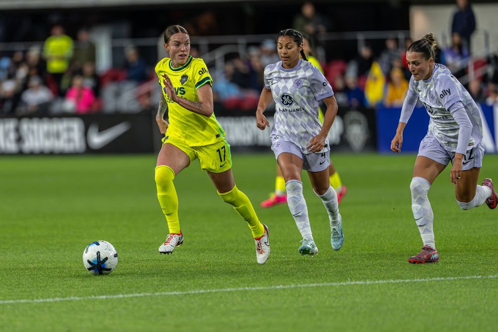 Female soccer players compete on the field, with a player in yellow dribbling the ball past opponents in white and gray uniforms.