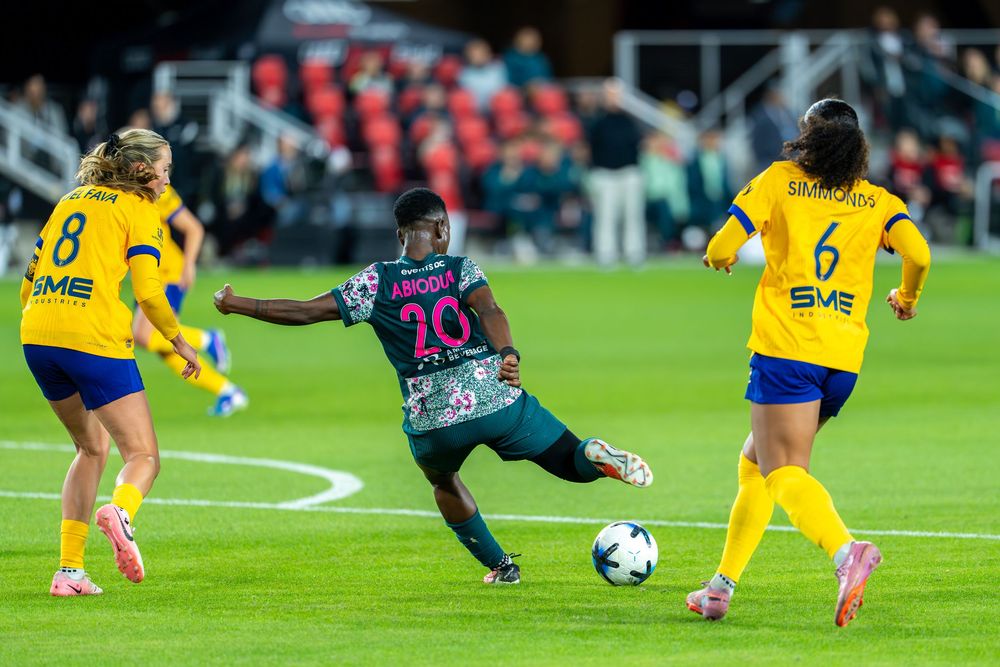 A soccer player in dark jersey battles for the ball against opponents in yellow uniforms during a women's football match.