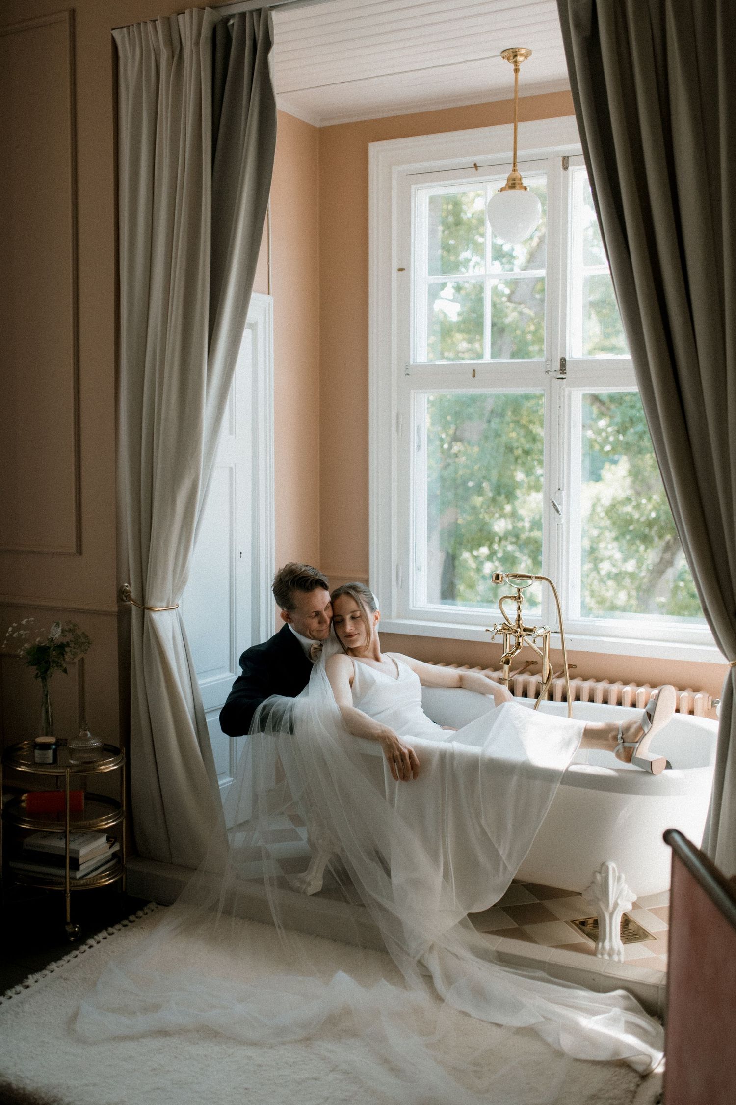 An artistic wedding photo showing a couple sitting in a bathtub by the window in an elegant wedding suite in Billnäs Gård, Finland.