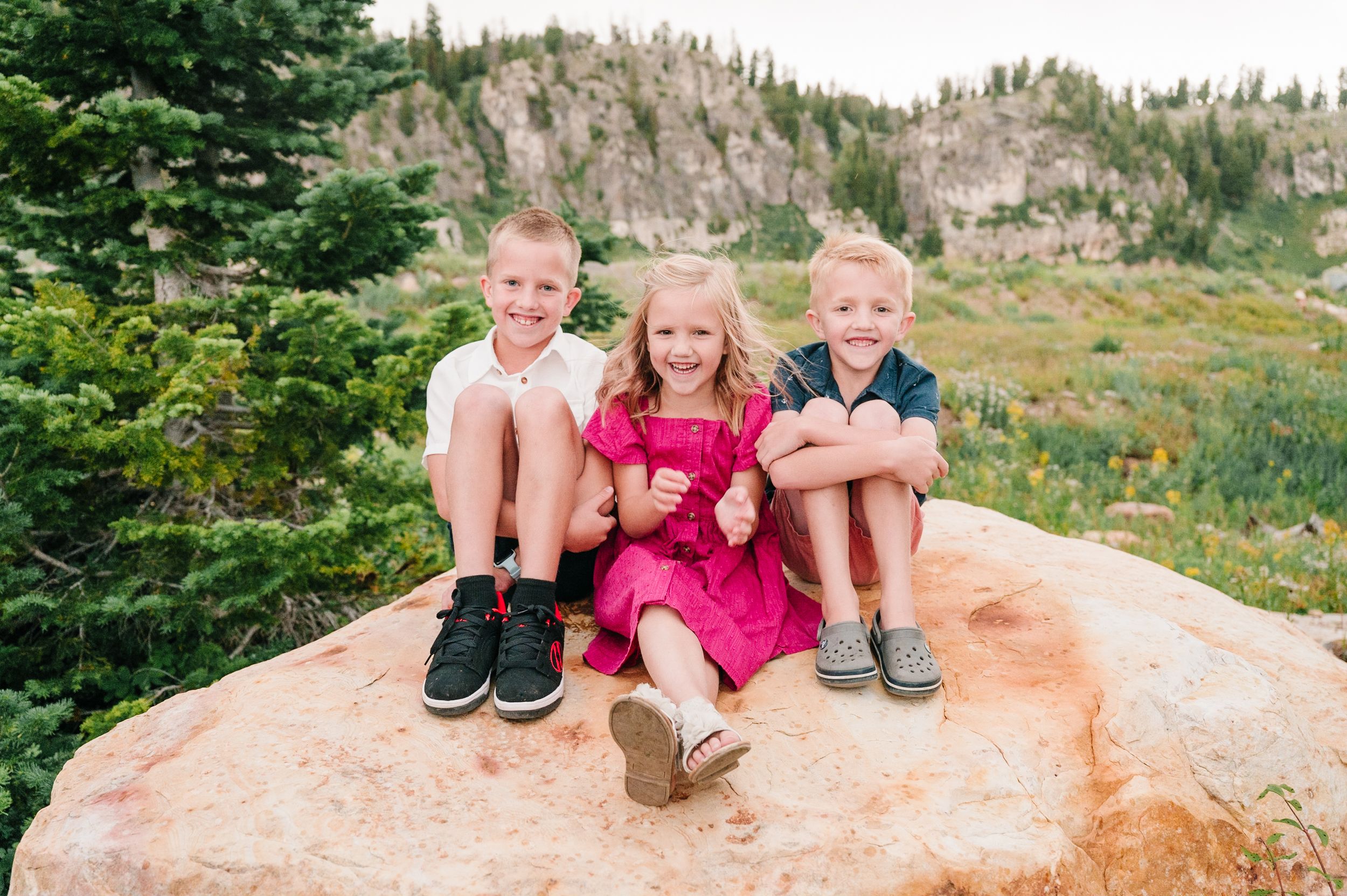 Three children sit together on large rock formation in mountain landscape.