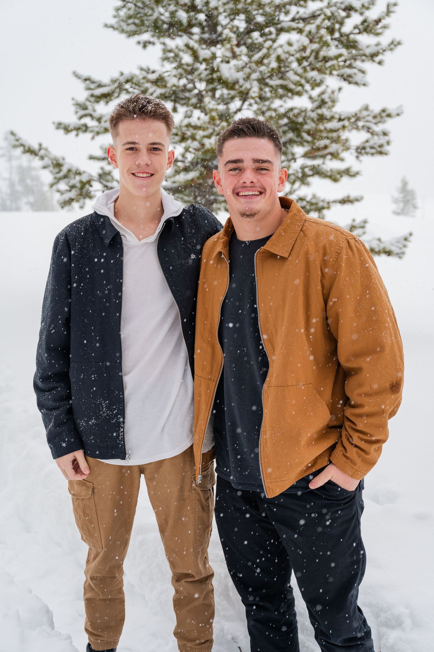 Two friends standing together in snow with evergreen trees behind them.