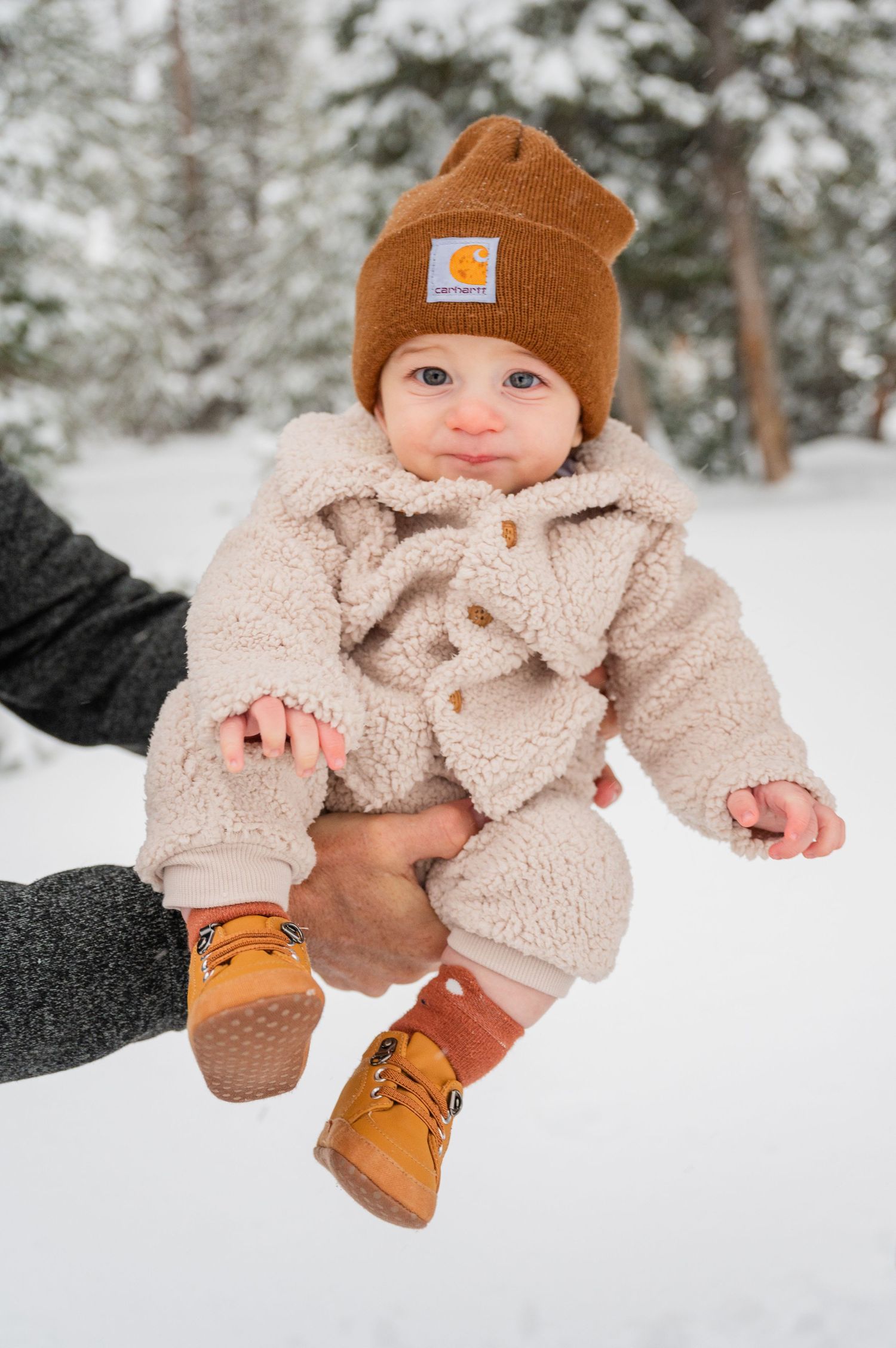 Baby wearing brown beanie and sherpa coat sits in snow during winter photoshoot.