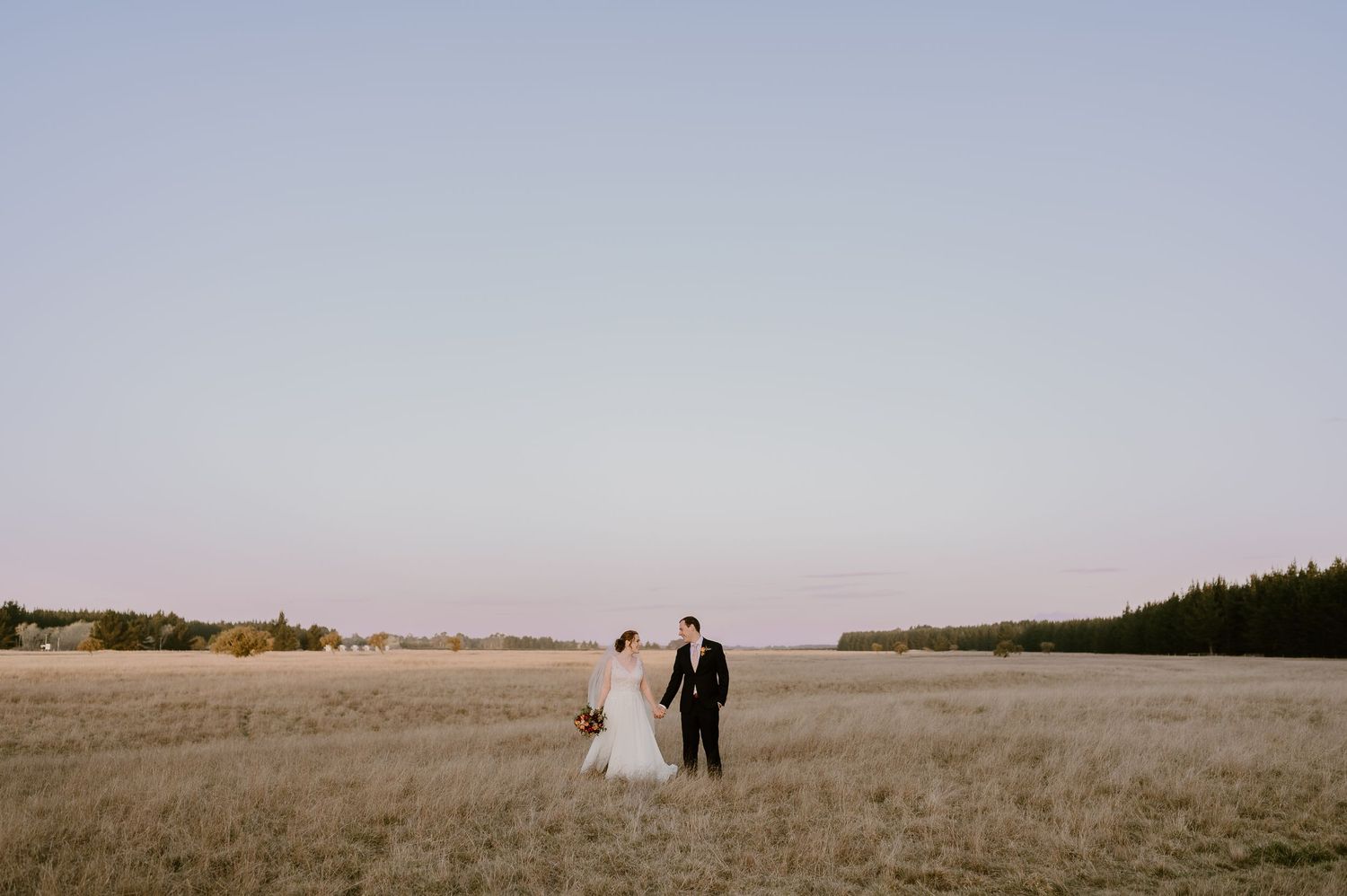 A couple walks hand in hand through a vast golden field at dusk with distant trees on the horizon.