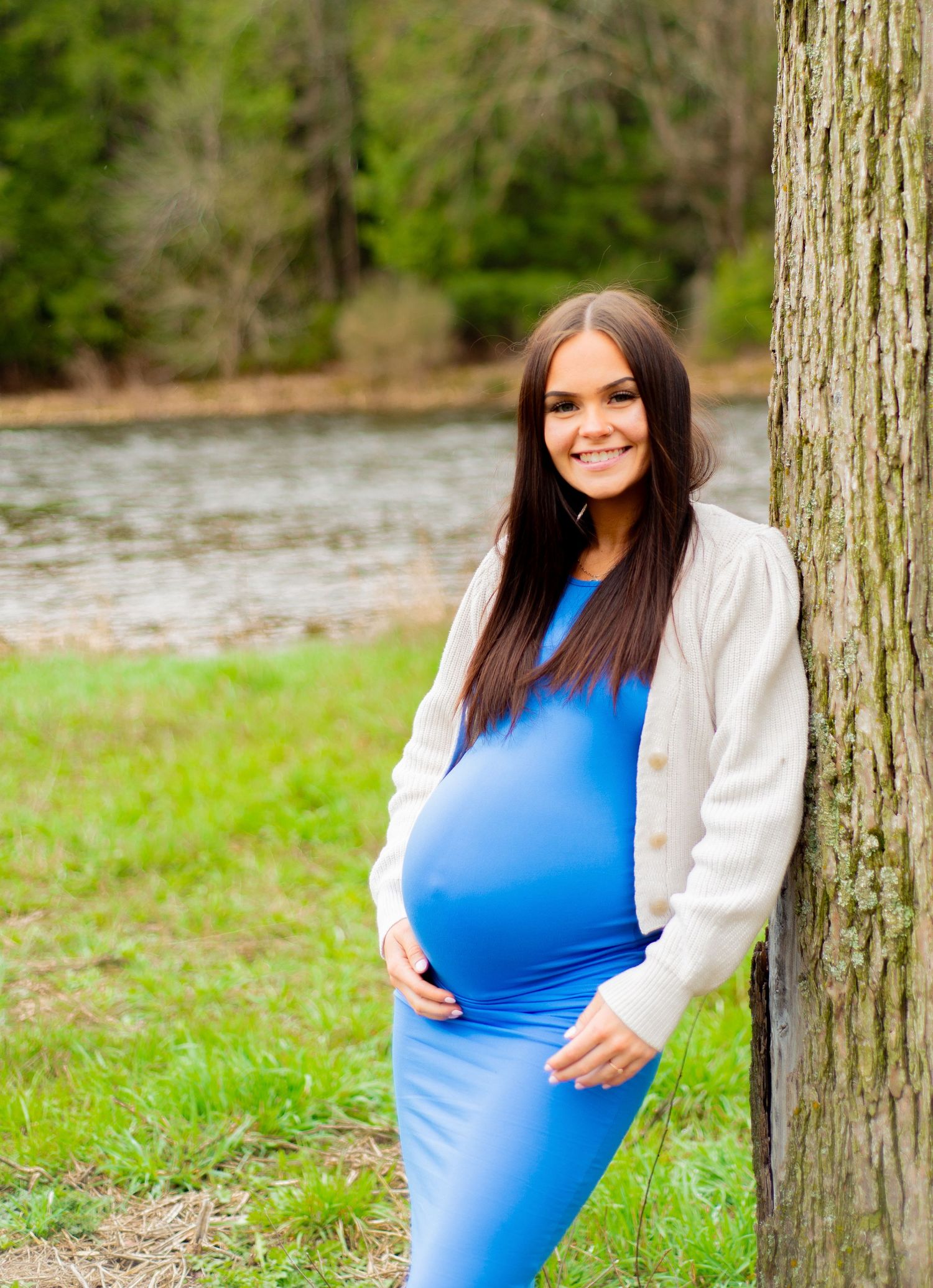 An expectant mother in a blue dress poses by a tree near a riverside.