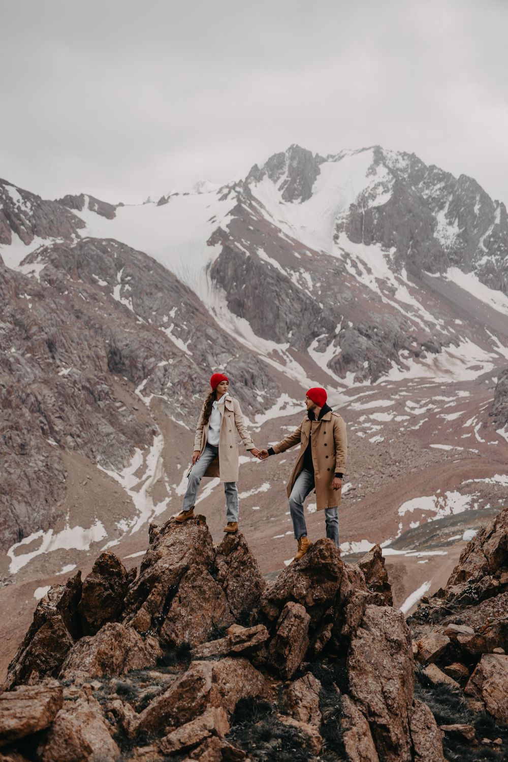 A couple holding hands, standing high on the rocks surrounded by towering mountains