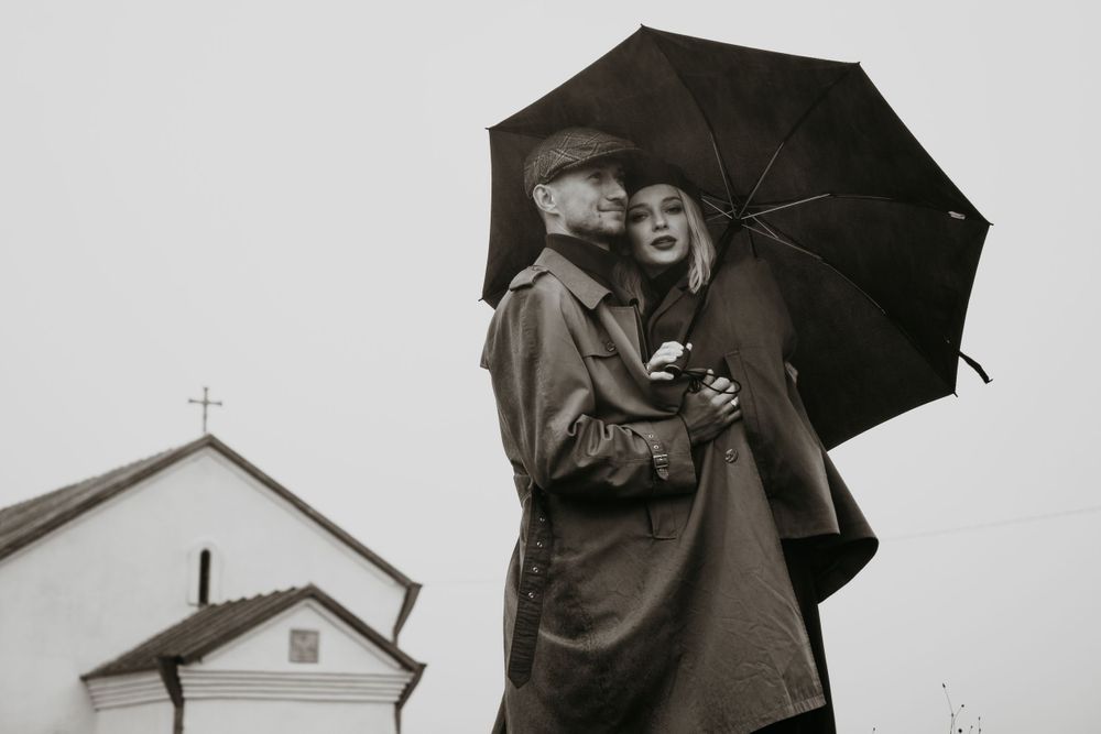 Black and white engagement photography of a couple hugging under the rain with an umbrella by the church