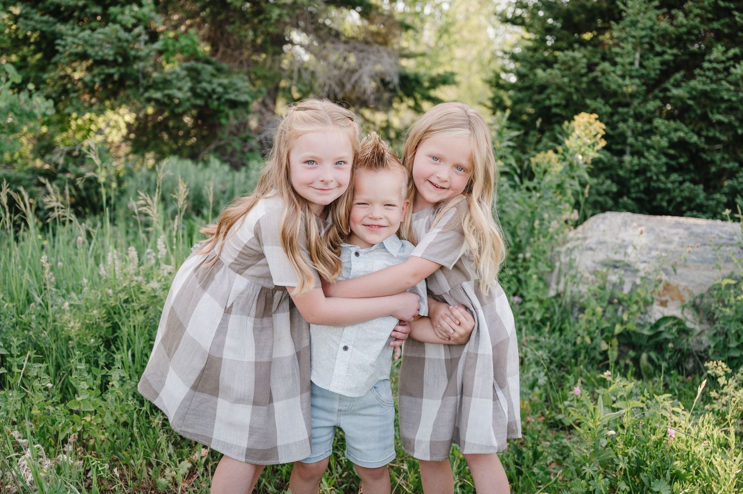 Three children in matching gray and white dresses embrace and smile together in a lush garden setting.