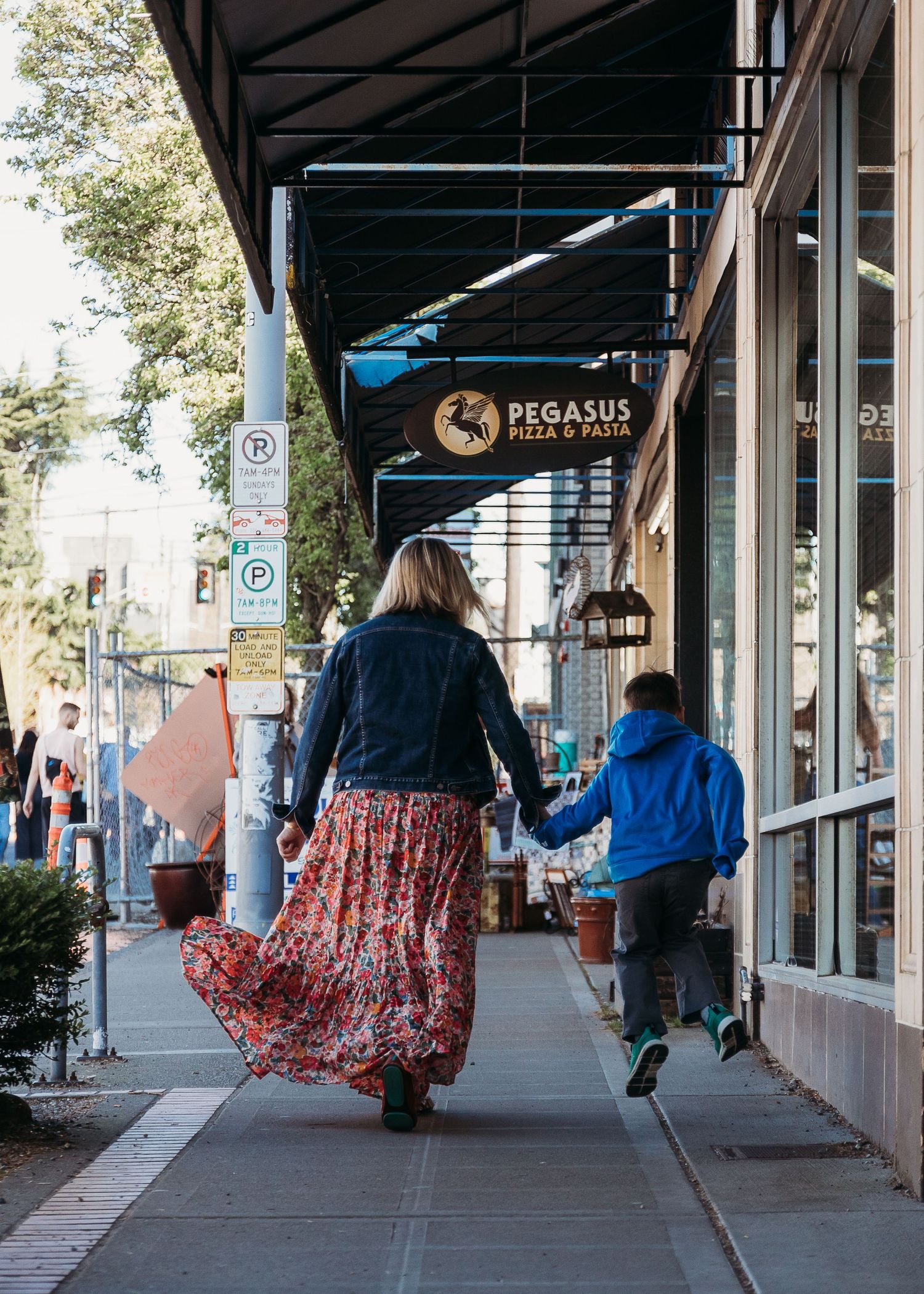 People walking under covered sidewalk past Pegasus Pizza shop sign in urban setting.