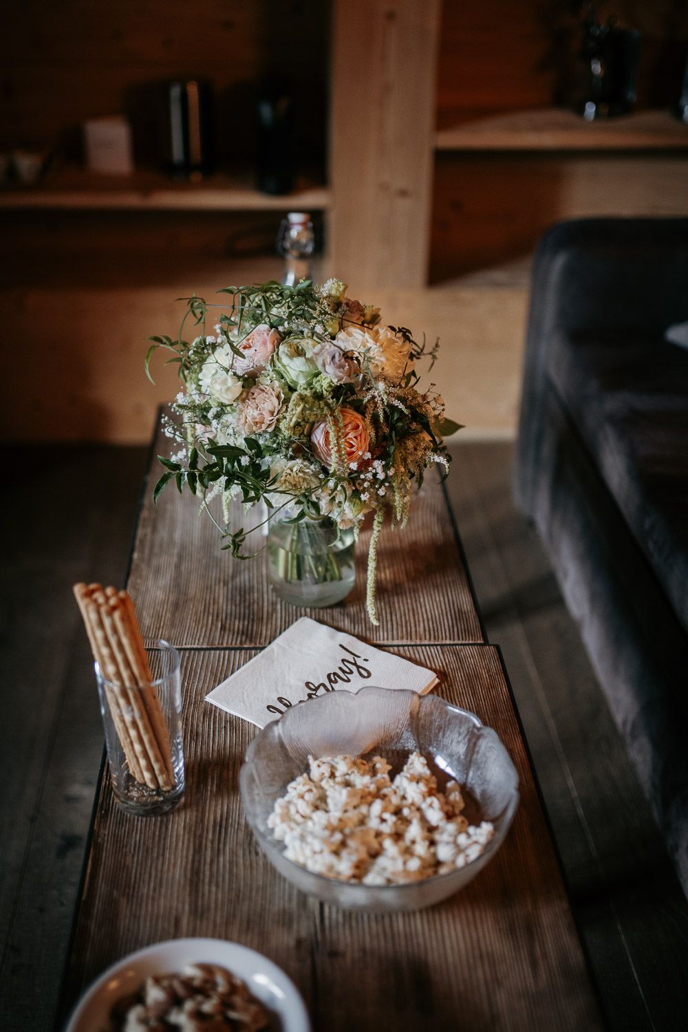Rustic wooden table displays bridal bouquet with orange and pink blooms beside popcorn and wedding card.