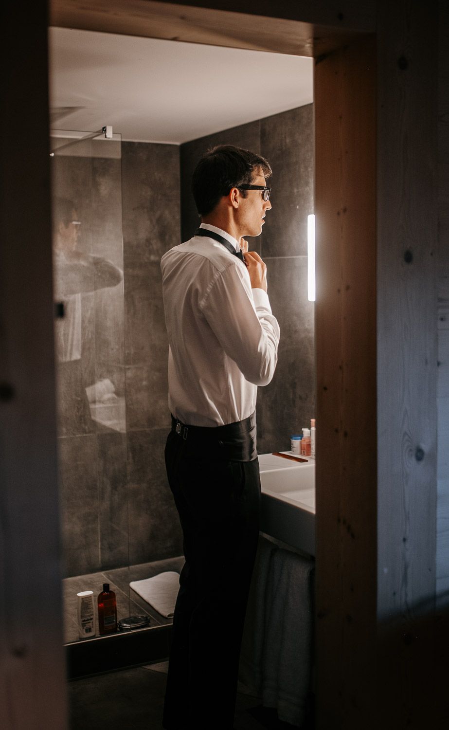 Groom in white shirt and suspenders adjusting bow tie in a modern bathroom mirror.
