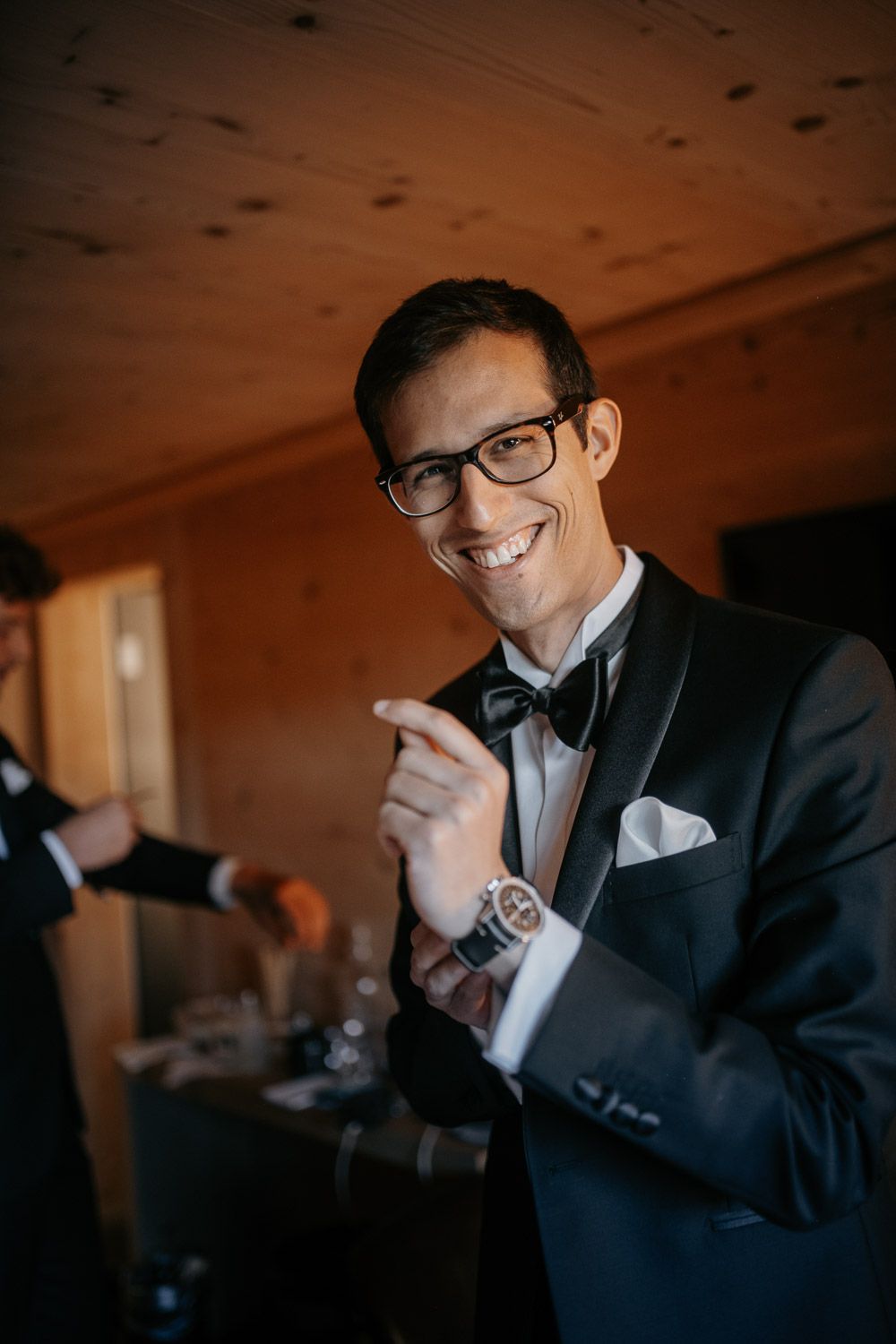 Smiling groom in black tuxedo and bow tie adjusting cufflinks in a wooden-ceiling venue.