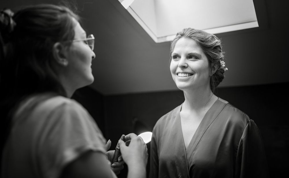 Black and white photo of bride smiling in robe while makeup artist works on her wedding day.