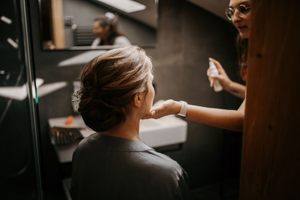 Hair stylist applying finishing spray to brides elegant updo with pearl accessory in bathroom.
